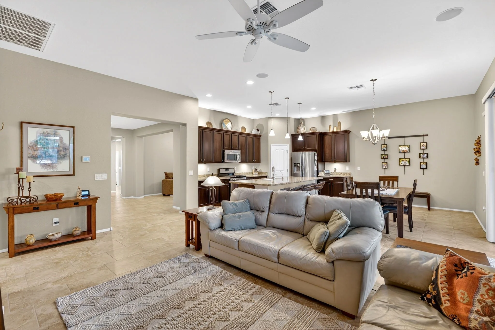 Open-concept living room and kitchen with a beige sofa, wooden coffee table, and a kitchen with dark wood cabinets and stainless steel appliances. Ceiling fan and pendant lighting are visible.