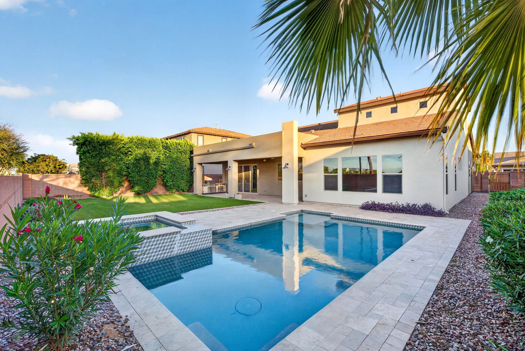 Residential backyard with swimming pool, surrounded by a well-maintained lawn, plants, and trees. The house has large windows and an outdoor grill area under a covered patio. A palm tree branch is visible in the upper right corner.