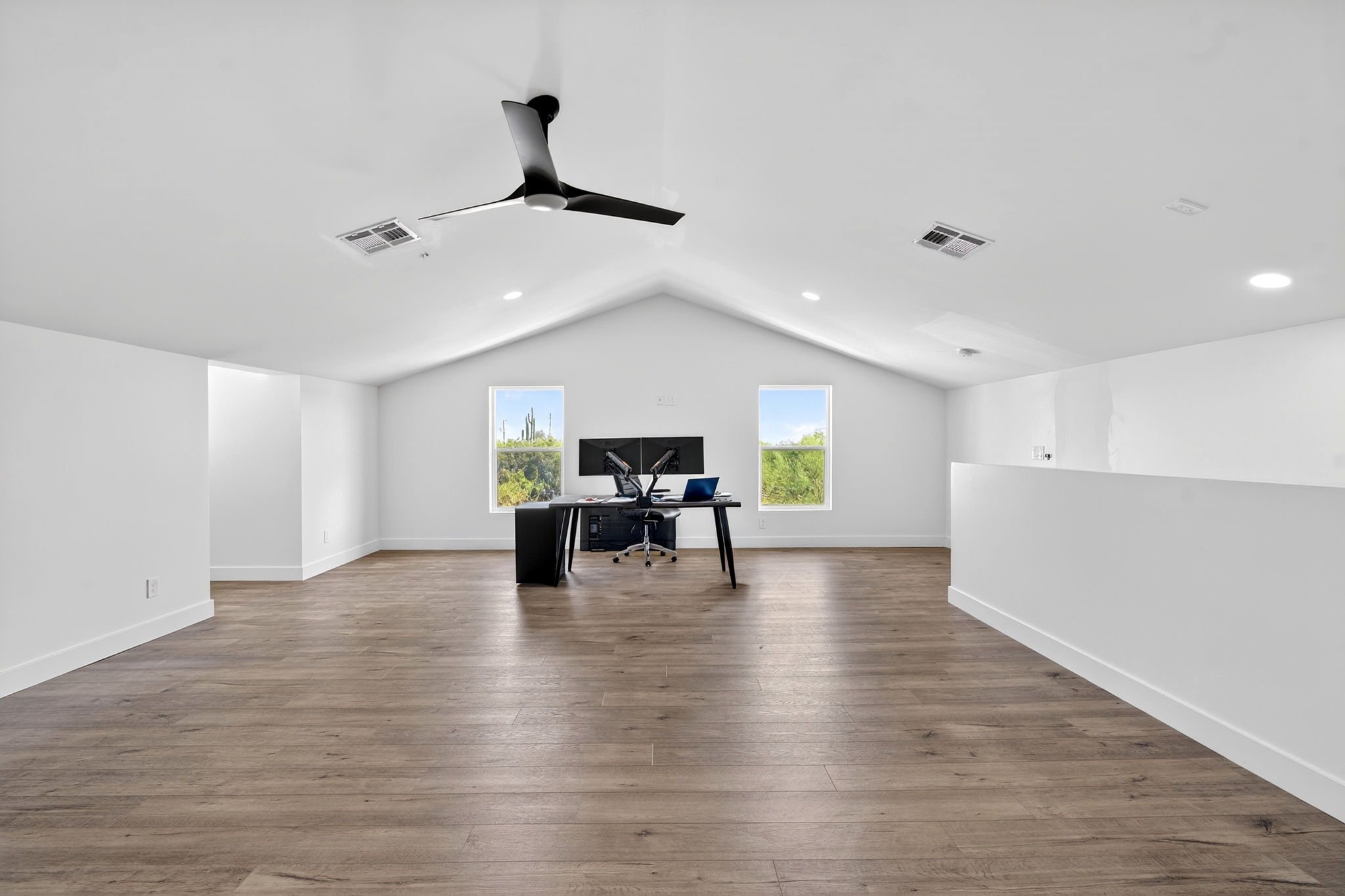 Empty in a modern, minimalist home office space with white walls, two windows, and wood flooring, containing a black desk, black office chair, and a large monitor.