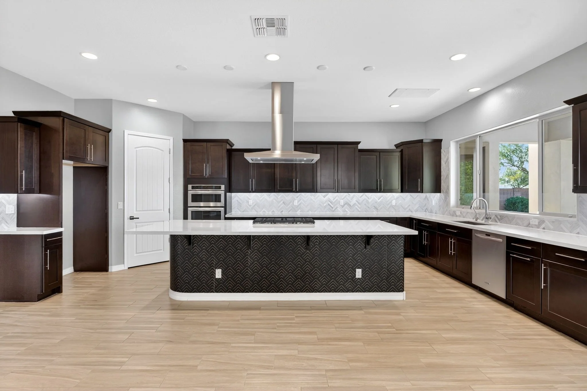 Modern kitchen with dark wood cabinets, a kitchen island with a patterned black facade, stainless steel range hood, and white marble backsplash, large window showing trees outside, and wood flooring.
