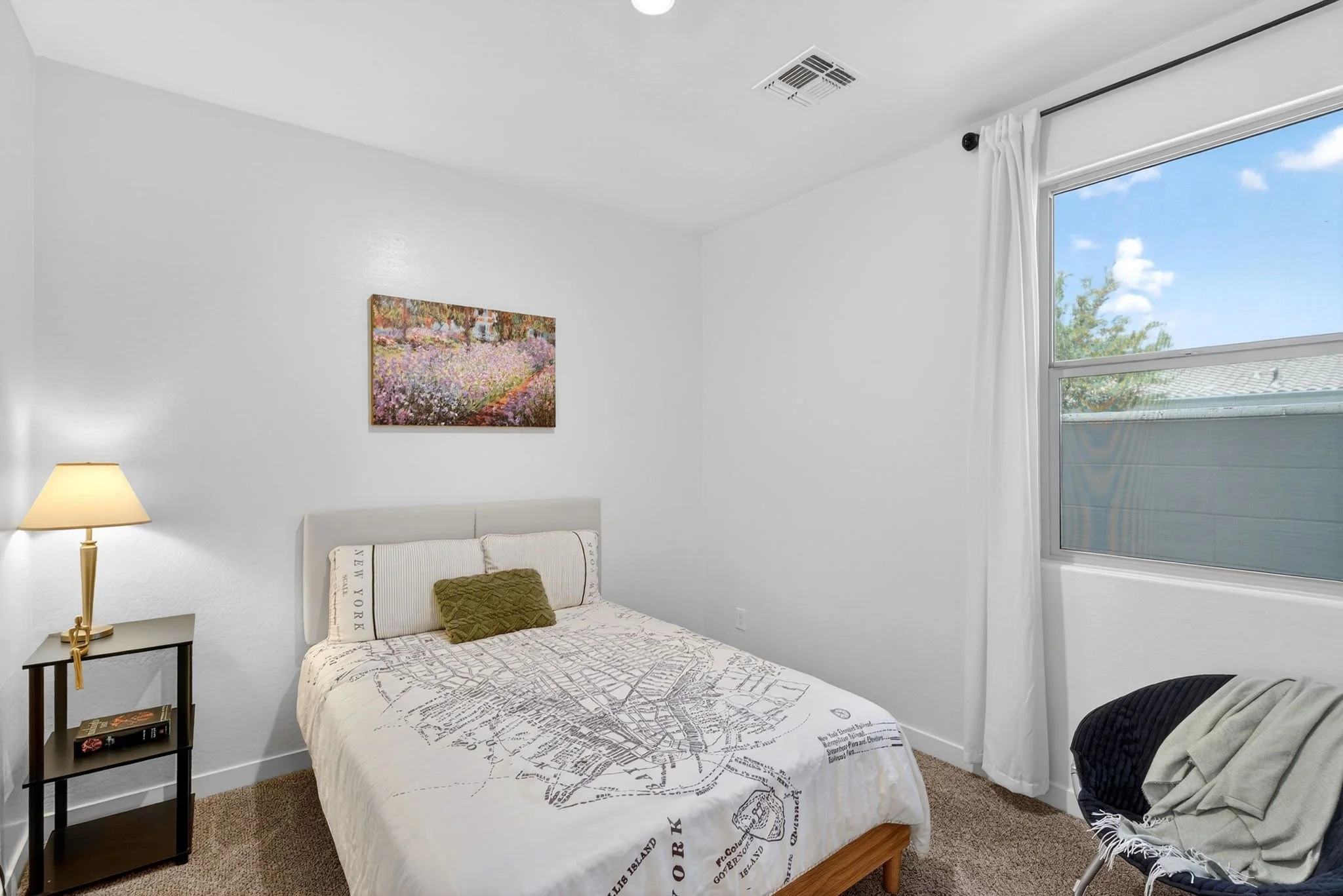 A bedroom with white walls, a window with a curtain, a bed with map-themed bedding, a side table with books, a lamp, a wall art of a garden, and a chair with a black cushion.