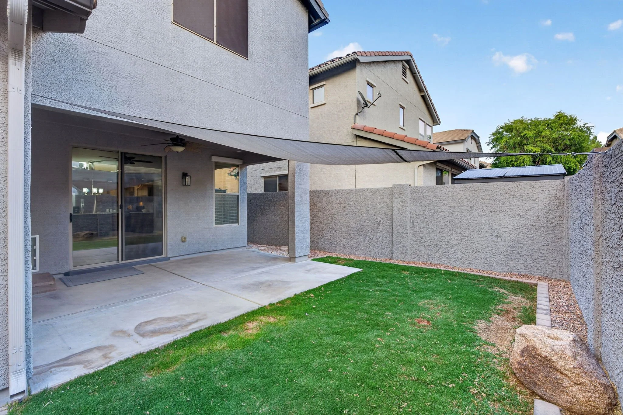 Backyard patio area with concrete flooring, sliding glass door, window, ceiling fan, covered area, adjacent green lawn, and tall wall with trees and neighboring houses visible beyond.