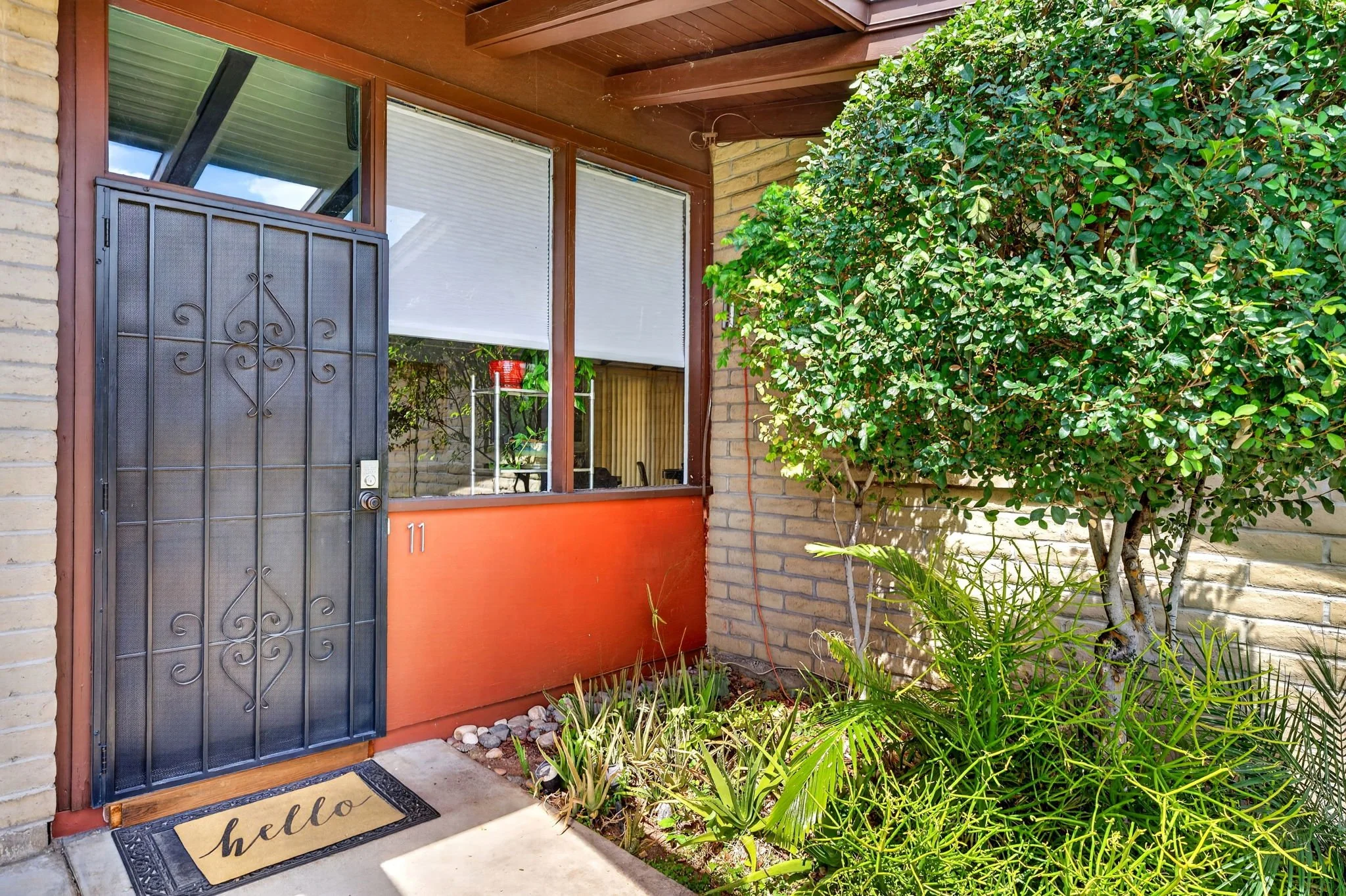 Front door of a house with a decorative black metal security door, a welcome mat with the word 'hello', and a small garden with green plants and bushes.