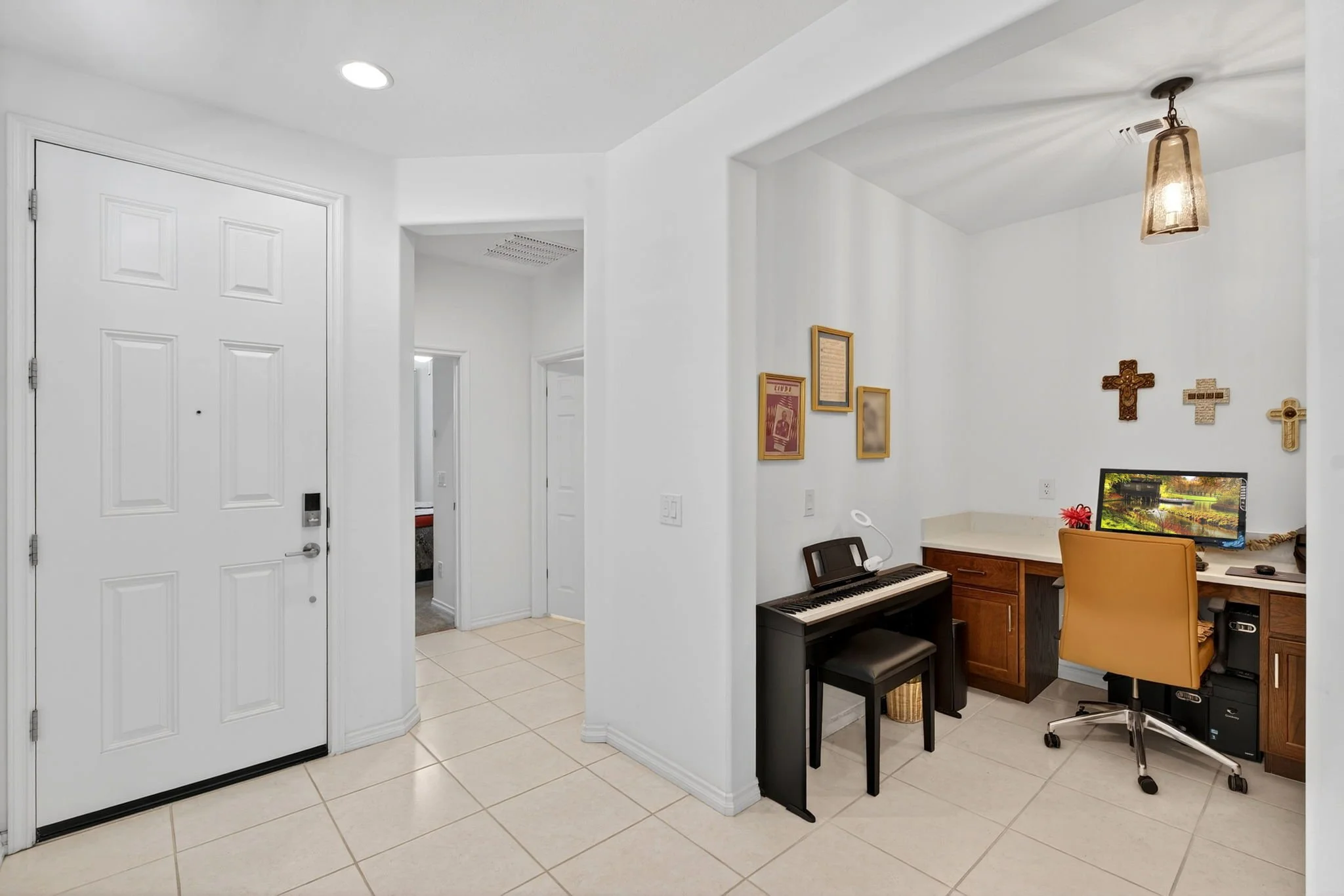 View of an entryway and a small home office corner with a digital piano, a desk with a computer monitor, and framed religious artwork on the wall, in a space with white walls and tiled flooring.
