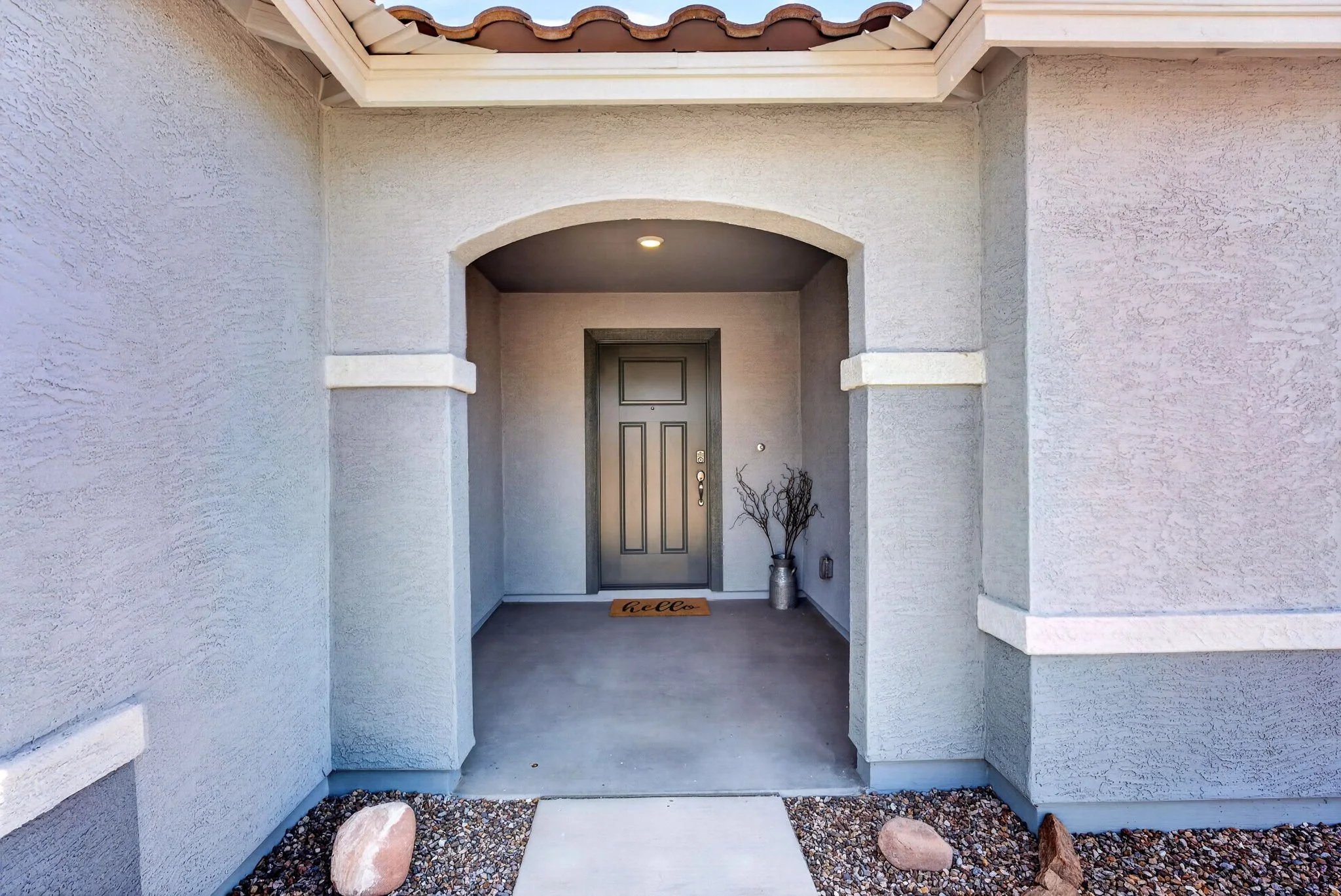 Front entry of a house with a covered porch, light-colored stucco walls, a black front door, a door mat that says "hello," a vase with decorative branches, and gravel with rocks on the ground.