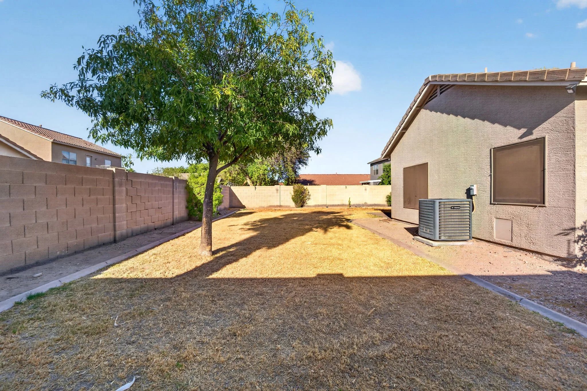 A backyard with a tree, a brick wall, a beige house with boarded-up windows, a central air conditioning unit, and a clear blue sky.