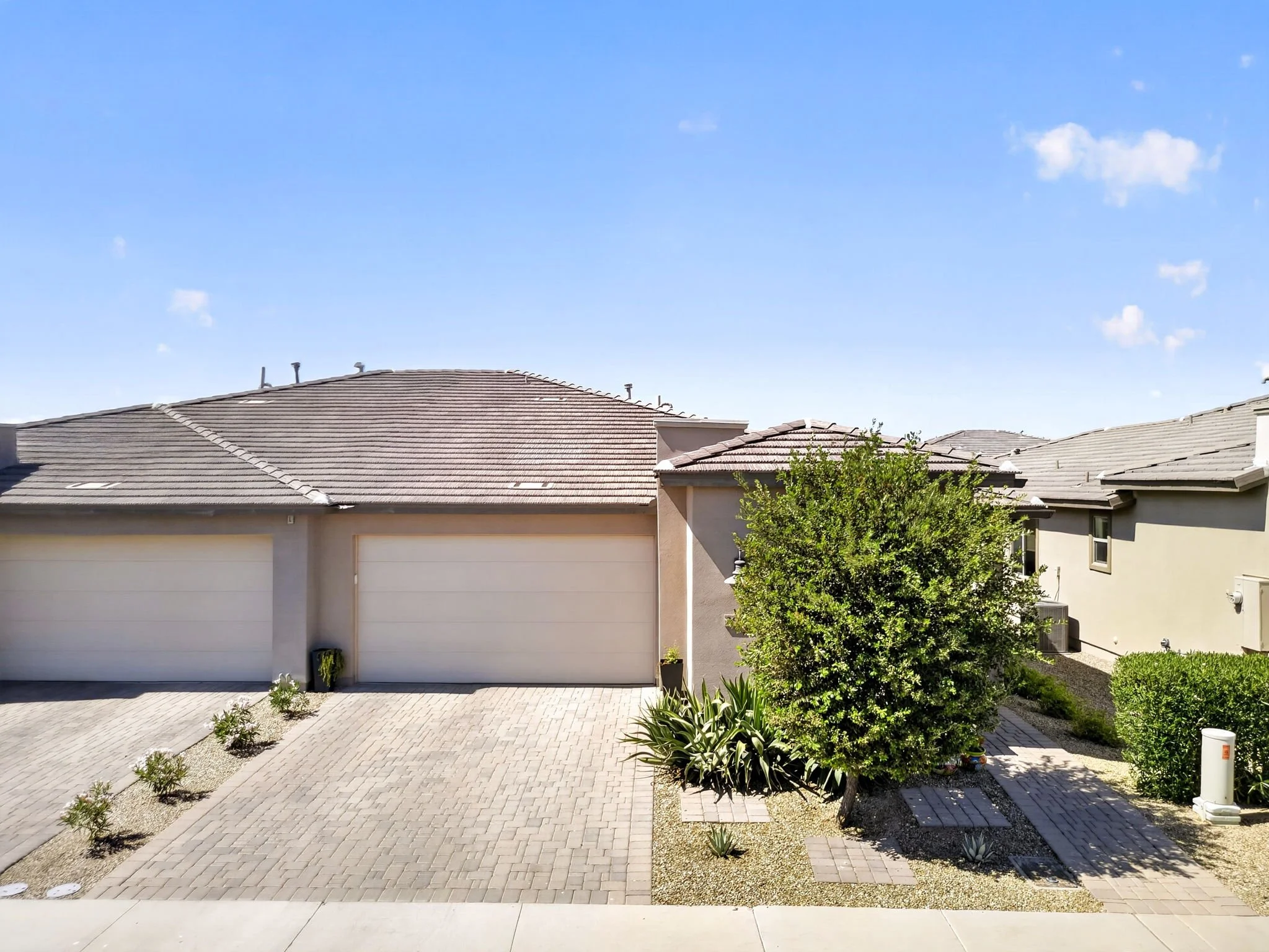 View of a suburban backyard with a paved patio, small shrubs, and a bush with green leaves, behind which are neighboring houses with tiled roofs, under a bright blue sky with a few white clouds.