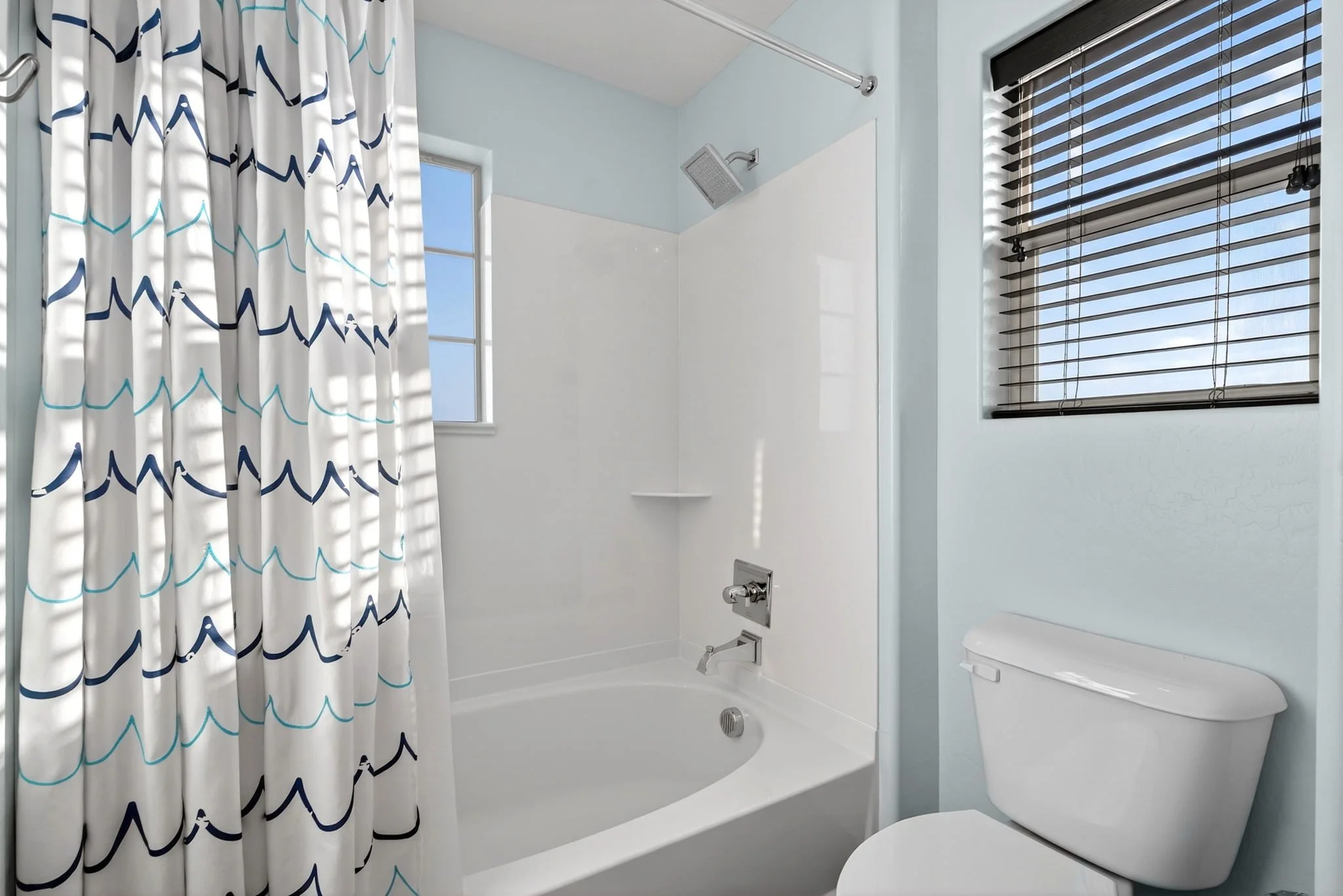 A clean, modern bathroom with a bathtub and shower combination. The bathtub has a showerhead and fixtures, and there is a shower curtain with a wave pattern partially covering the tub. The bathroom has a window with blinds and light blue walls.