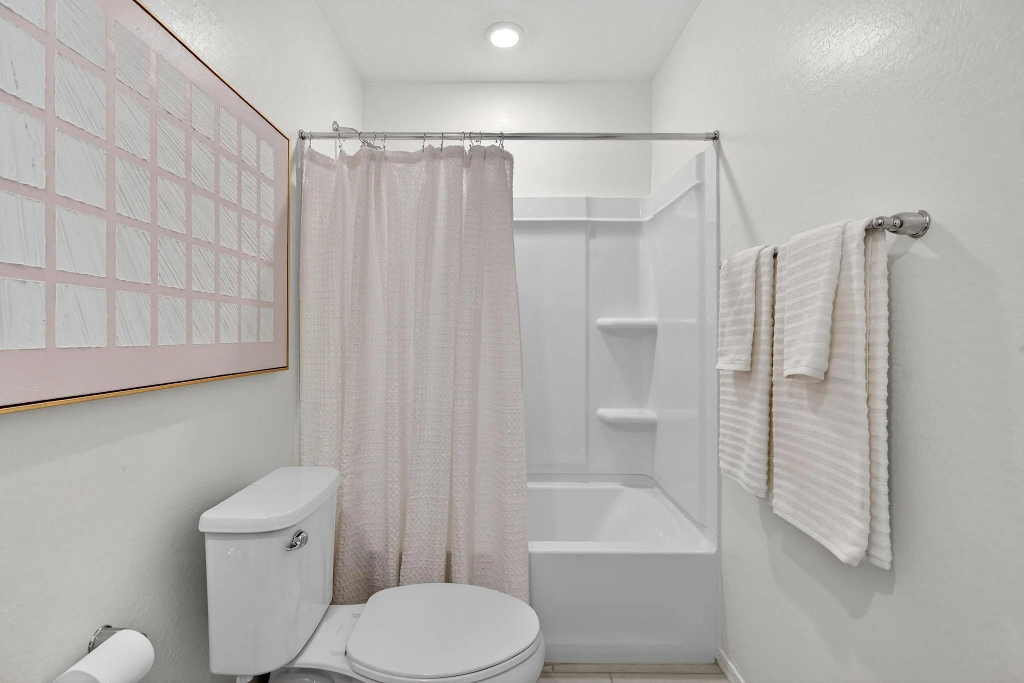 A small bathroom featuring a toilet, a shower with a pink shower curtain, towel racks with white towels, a wall with a framed artwork, and a white plastic shower surround.
