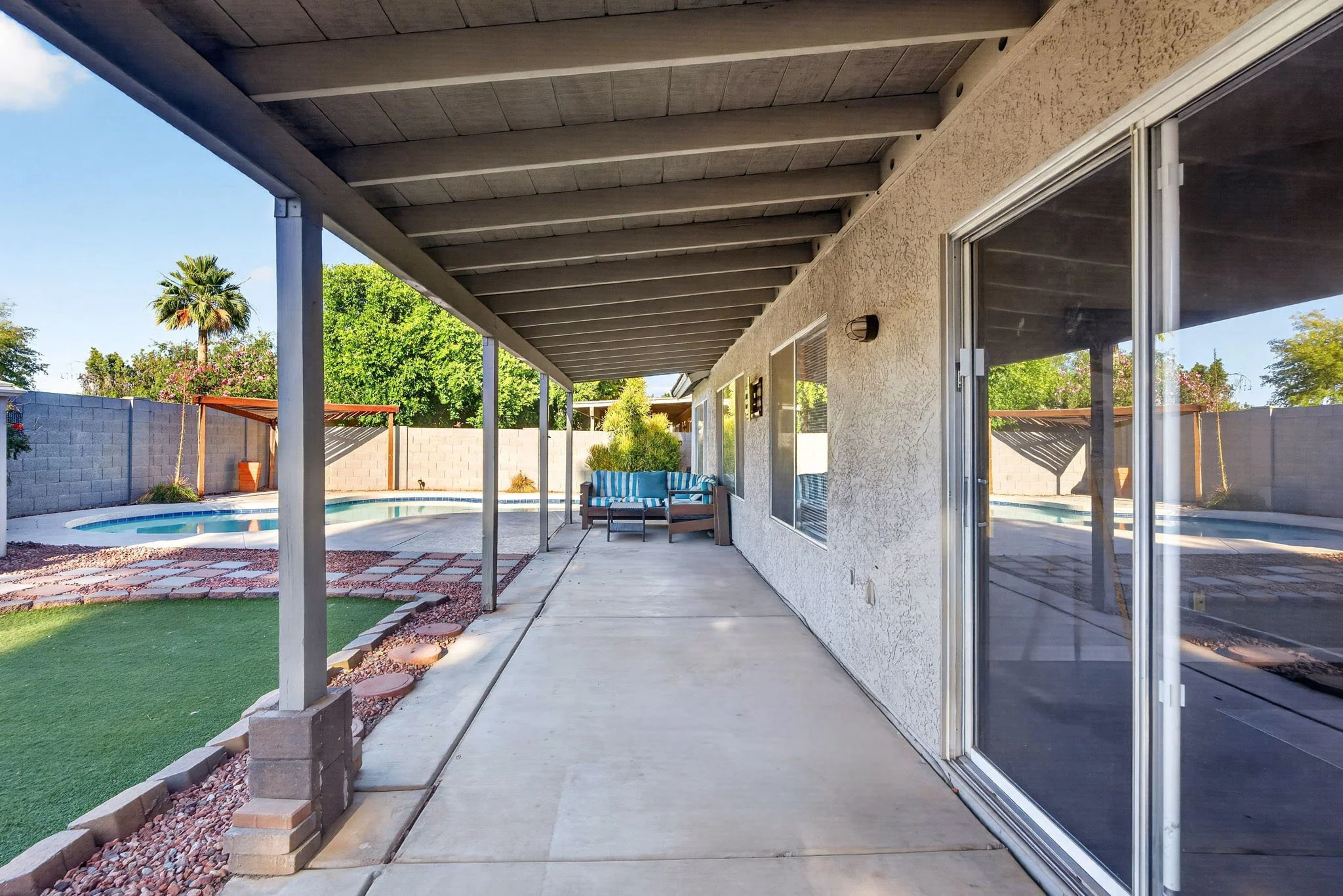Backyard patio with covered area, sliding glass door, outdoor seating, pool, and landscaped yard with trees.