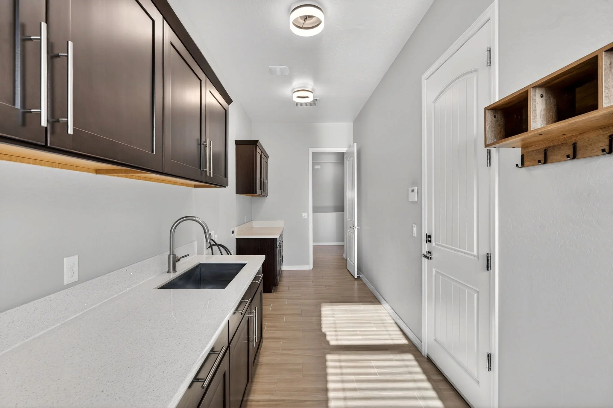 A modern kitchen with dark wood cabinets, white countertop, stainless steel sink, and light wood flooring. A white door with vertical panels is on the right, with a wooden wall hook rack above. The ceiling has two round light fixtures, and natural li