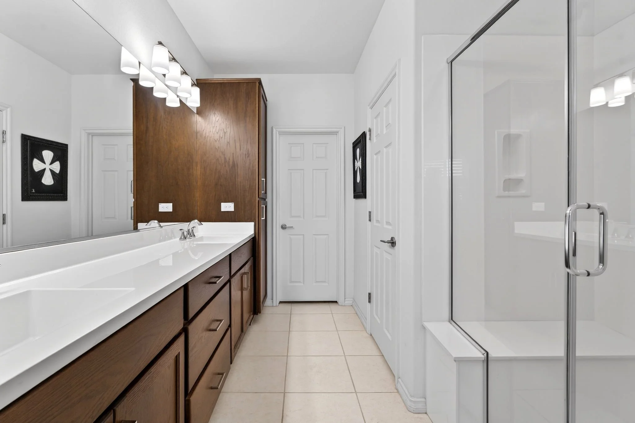 A bathroom with a white vanity with a dark wood cabinet, a large mirror, and a modern light fixture above. There is a white door in the background and a glass shower door on the right side of the image.
