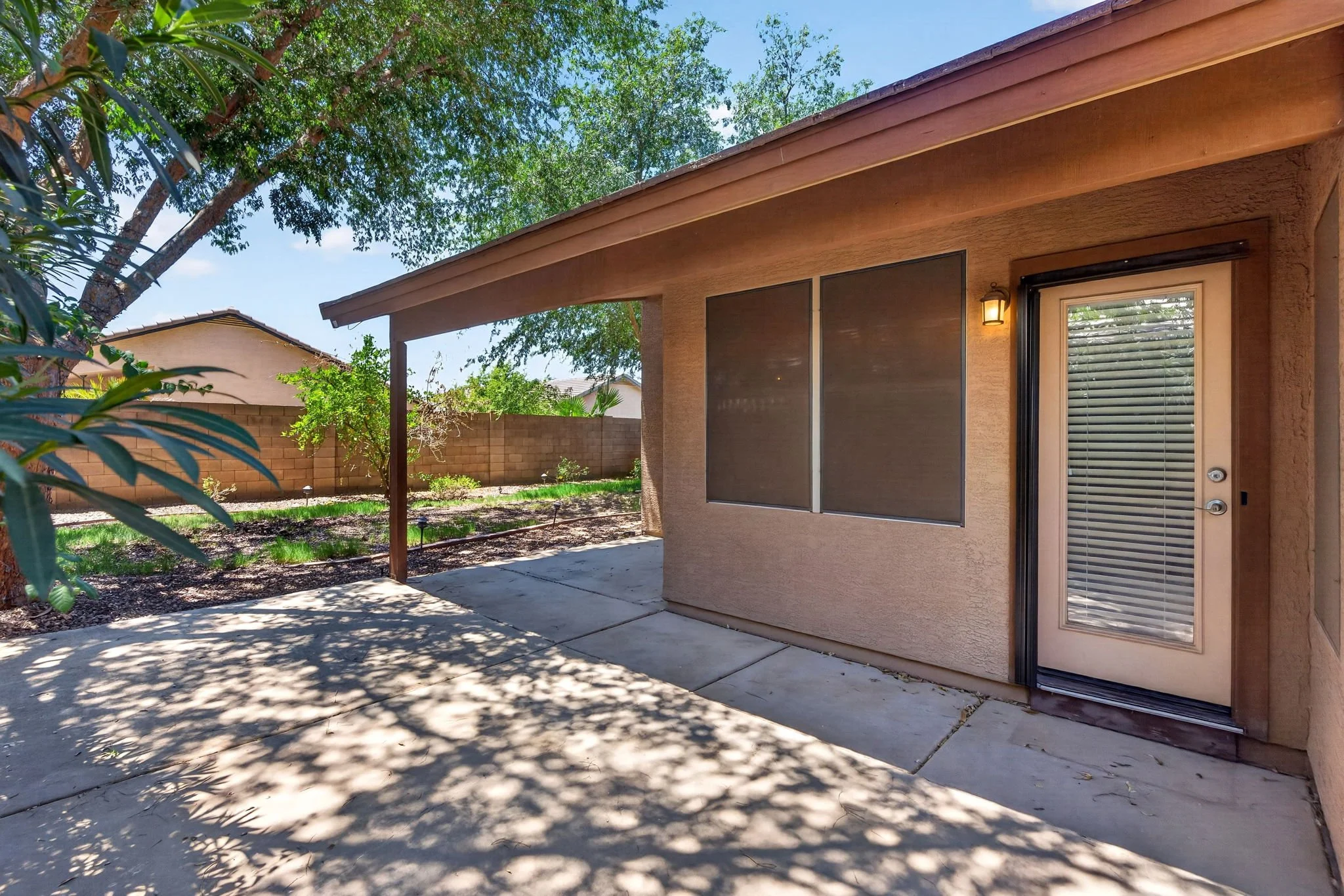 Backyard patio area with concrete flooring, shaded by a tree, with a brick wall in the background and a view of neighboring house. Part of the house's exterior wall with window, door, and outdoor light fixture are visible.