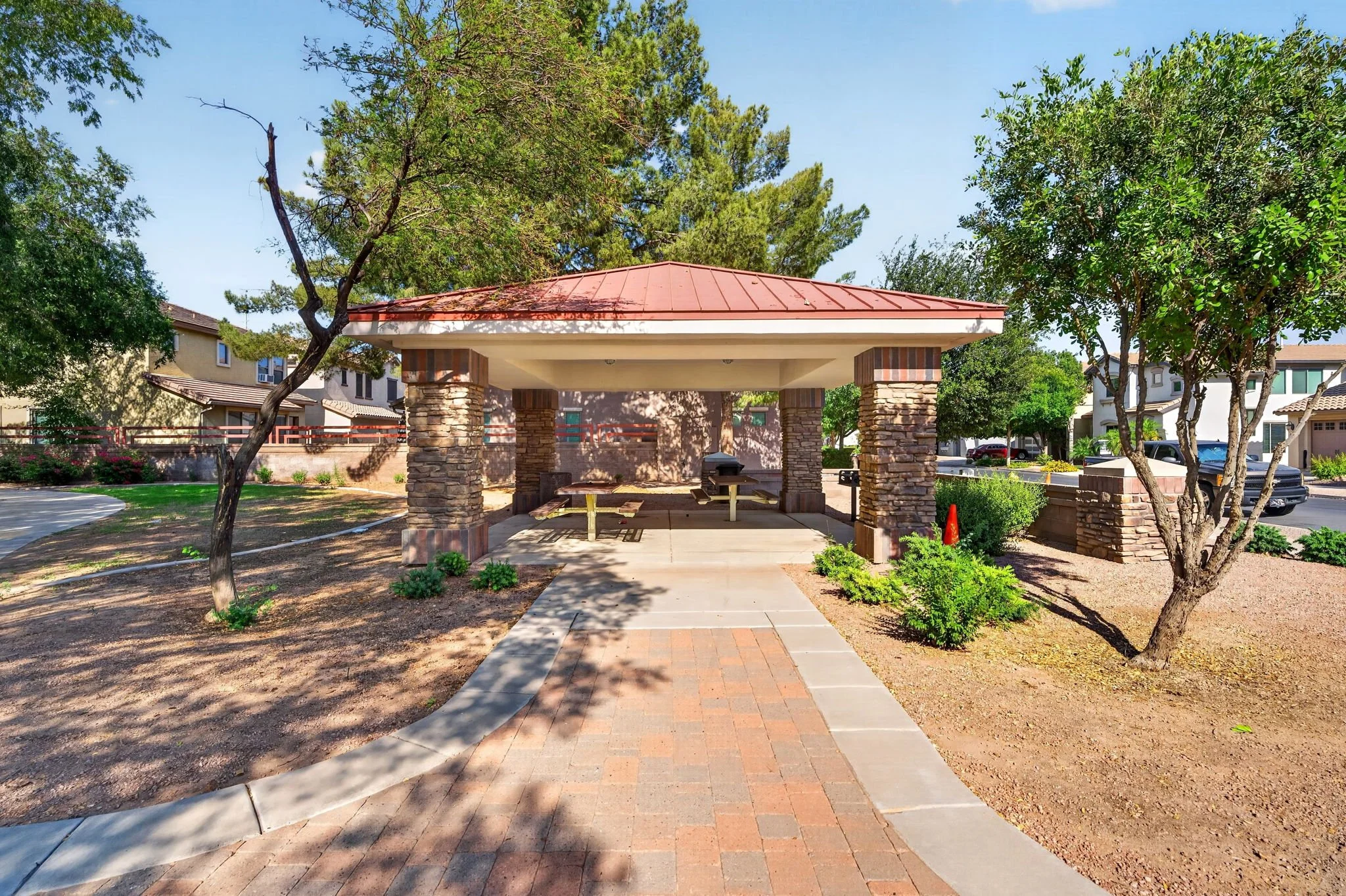 A park shelter with brick pillars and a red roof, surrounded by trees, with a concrete path leading to it and houses in the background.