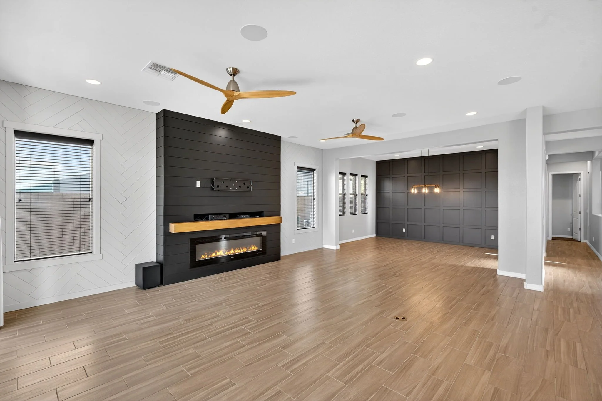 Modern living room with wood-look tile flooring, white walls, black accent wall with an electric fireplace, multiple windows with blinds, ceiling fans, and recessed lighting.