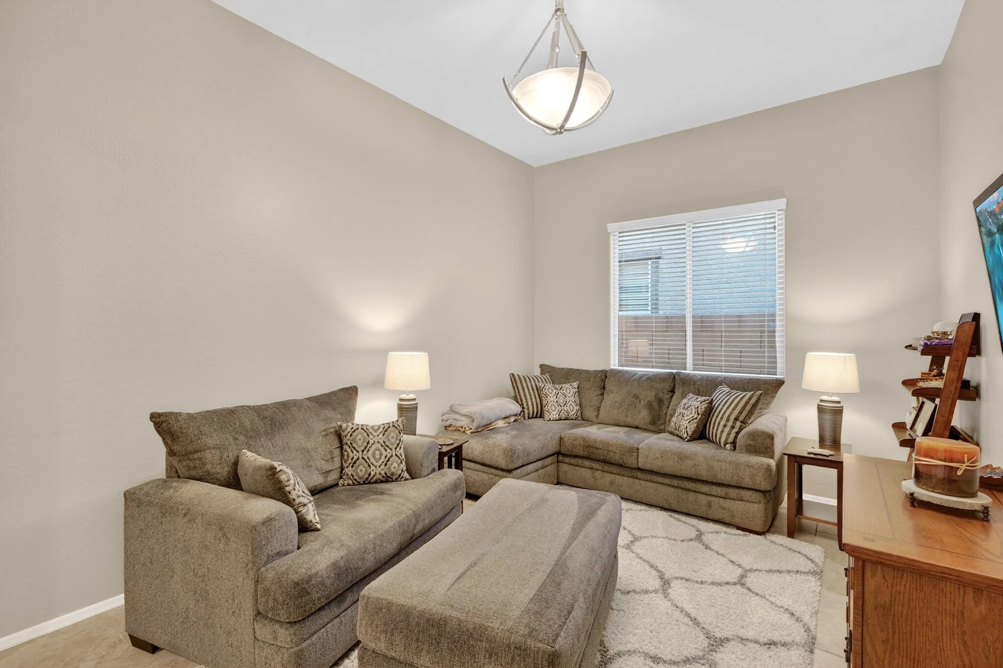 Living room with beige sofas, patterned pillows, lamps, window with blinds, and wall-mounted TV.