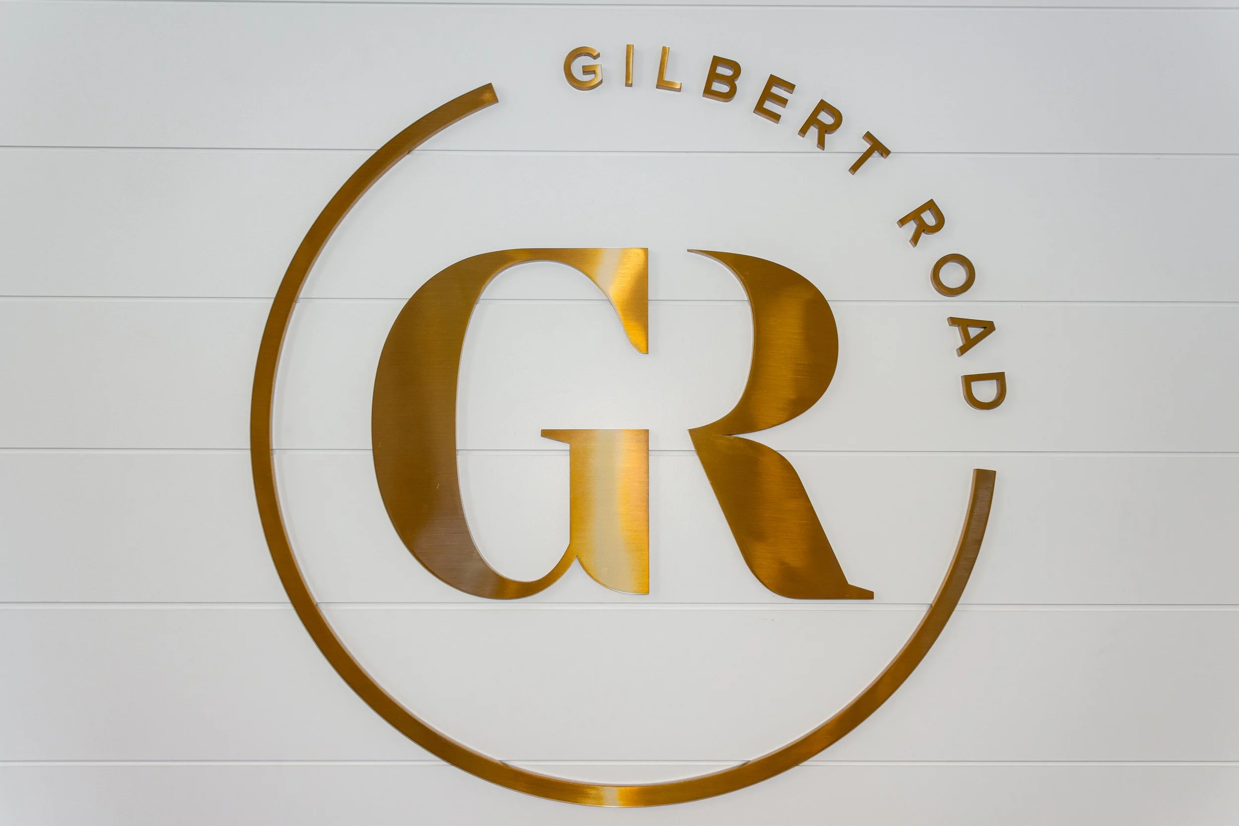 Golden logo of Giltner on a white wall with horizontal paneling, featuring a stylized 'G' enclosed within a circular outline and the text 'GILBERT ROAD' curved above.