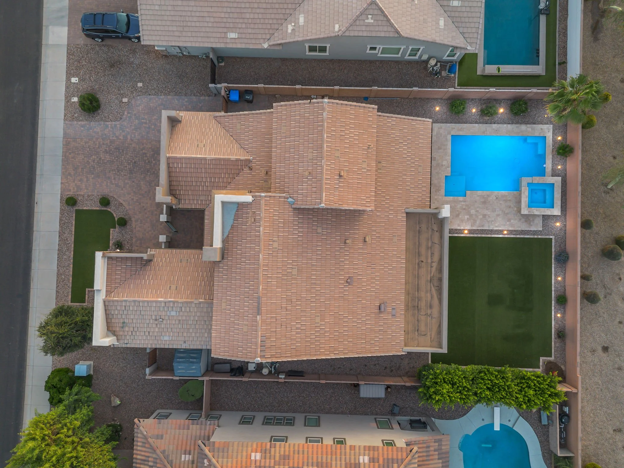 An aerial view of a backyard featuring a swimming pool with surrounding paved stone area, a shade structure, small green lawn, trees, a gravel pathway, and neighboring houses.