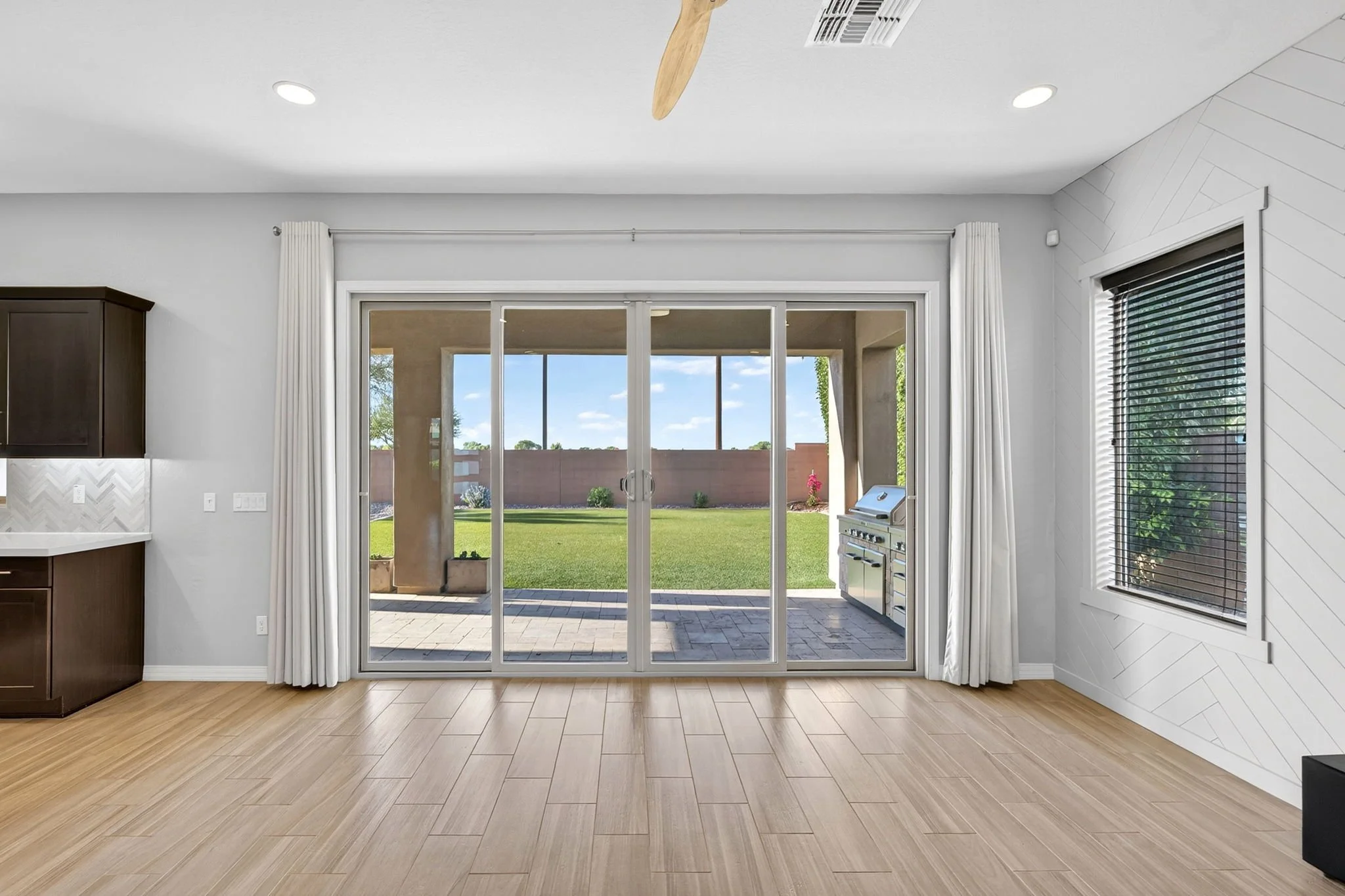 View of a modern kitchen with a sliding glass door leading to a backyard with a patio, grassy lawn, and a wooden fence. Kitchen features dark cabinetry, light-colored walls, a window with blinds, and hardwood floors.