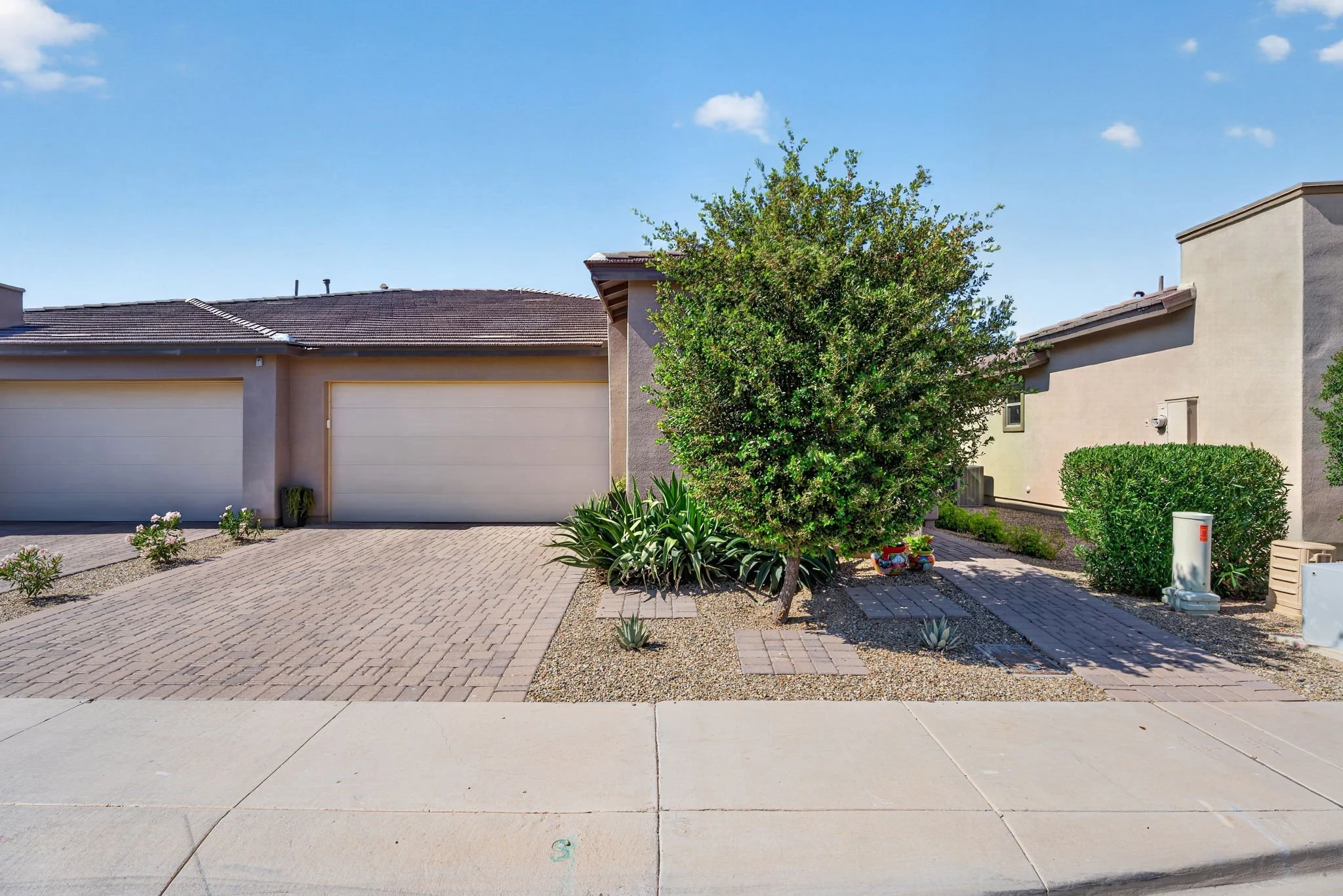 Front yard of a house with a garage, a large leafy tree, small bushes, and a paved walkway under a blue sky with a few clouds.
