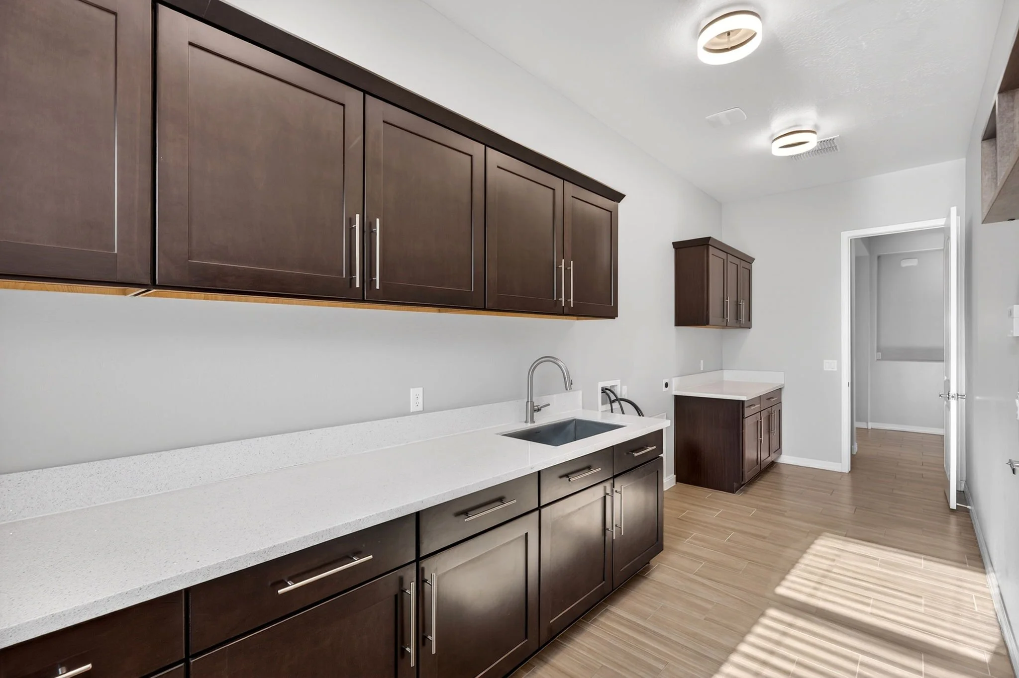 Modern kitchen with white countertops, dark brown cabinets, a stainless steel sink, and hardwood flooring, illuminated by ceiling lights.