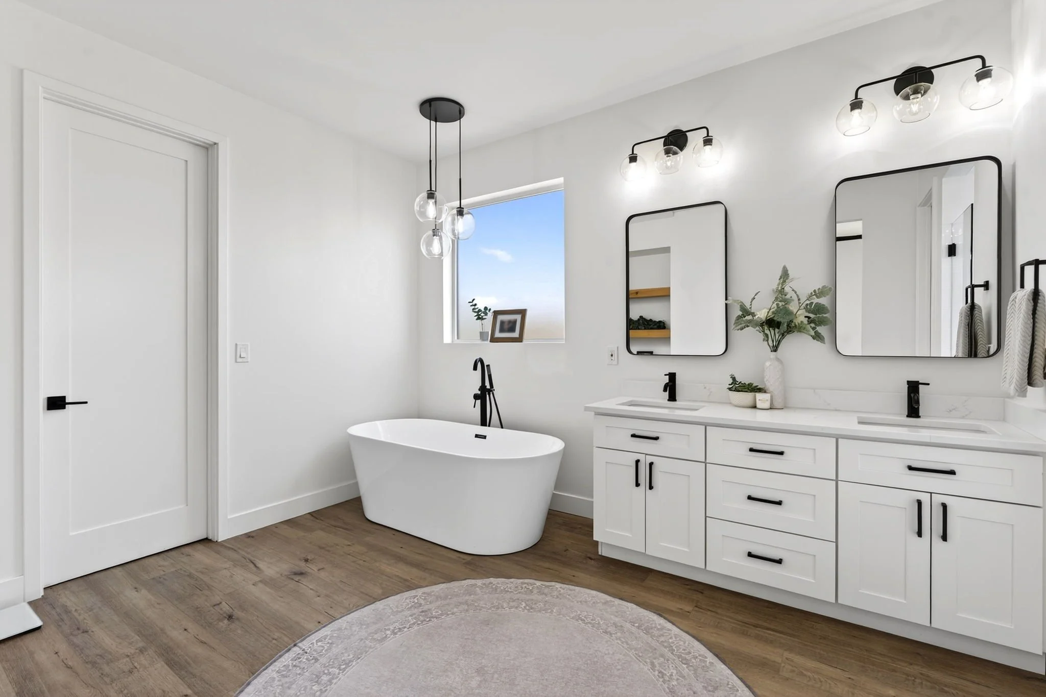 Modern bathroom with a white freestanding bathtub, white vanity with black hardware, two black-framed mirrors, and a window with a small plant and picture on the sill. There are two light fixtures, one with multiple bulbs and the other with fewer, cl