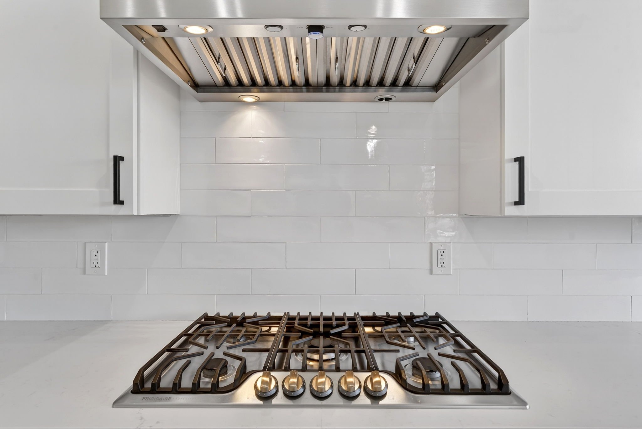 View of a kitchen stove with a gas cooktop and a vent hood overhead against a white tiled wall.