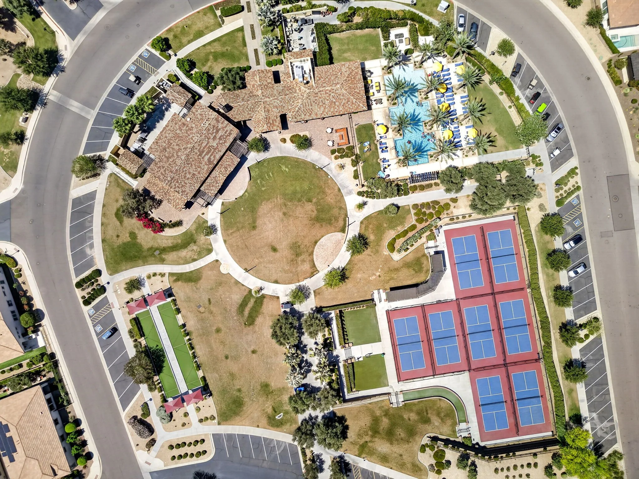 An aerial view of a residential community featuring a large swimming pool with yellow and blue umbrellas, palm trees, tennis courts, and parking lots. The area is surrounded by roads and green lawns.