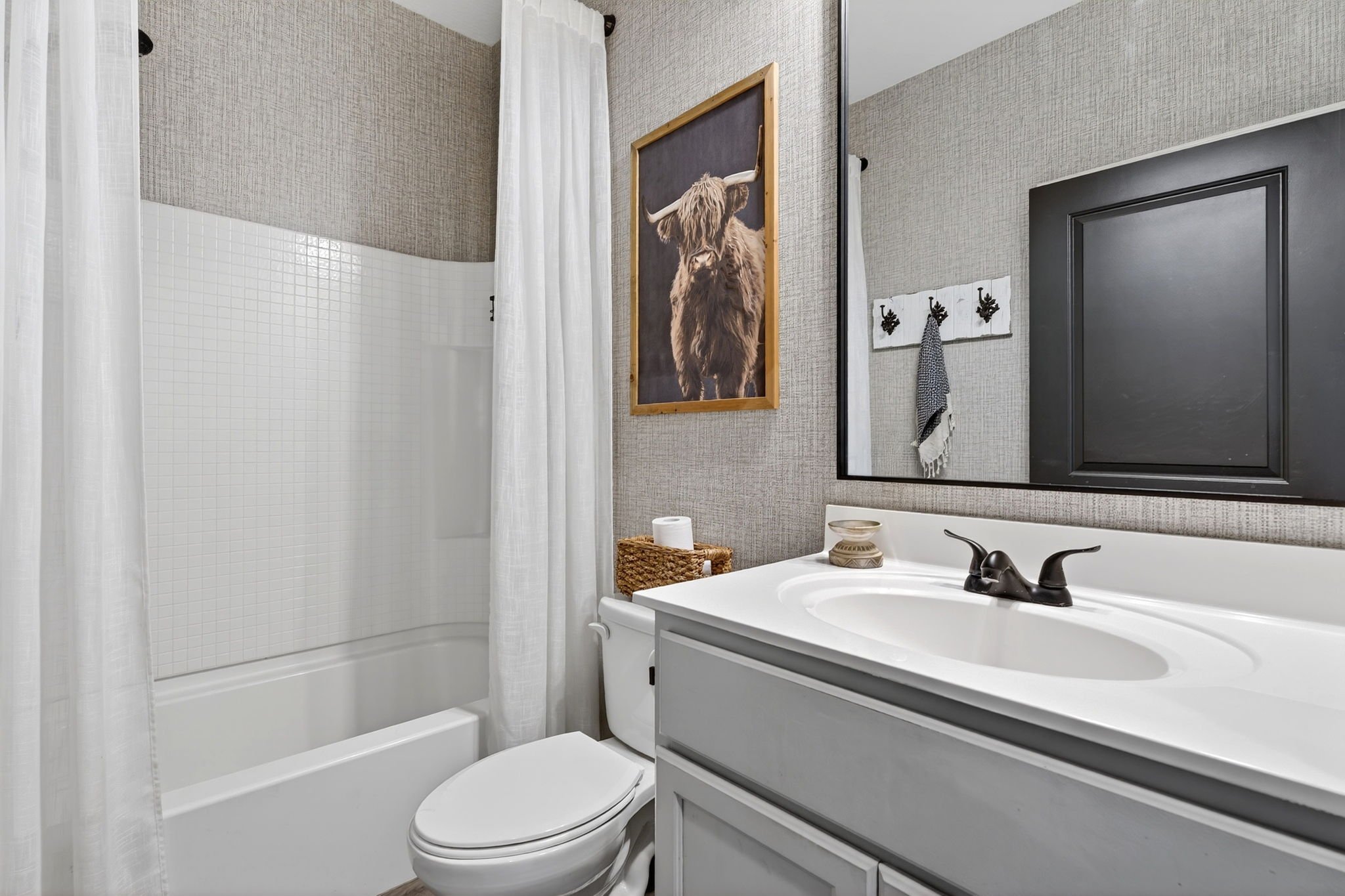 Bathroom with a shower, white curtains, wall art of a Highland cow, mirror, towel hooks, a black framed mirror, a beige basket, and a white vanity with a black faucet.