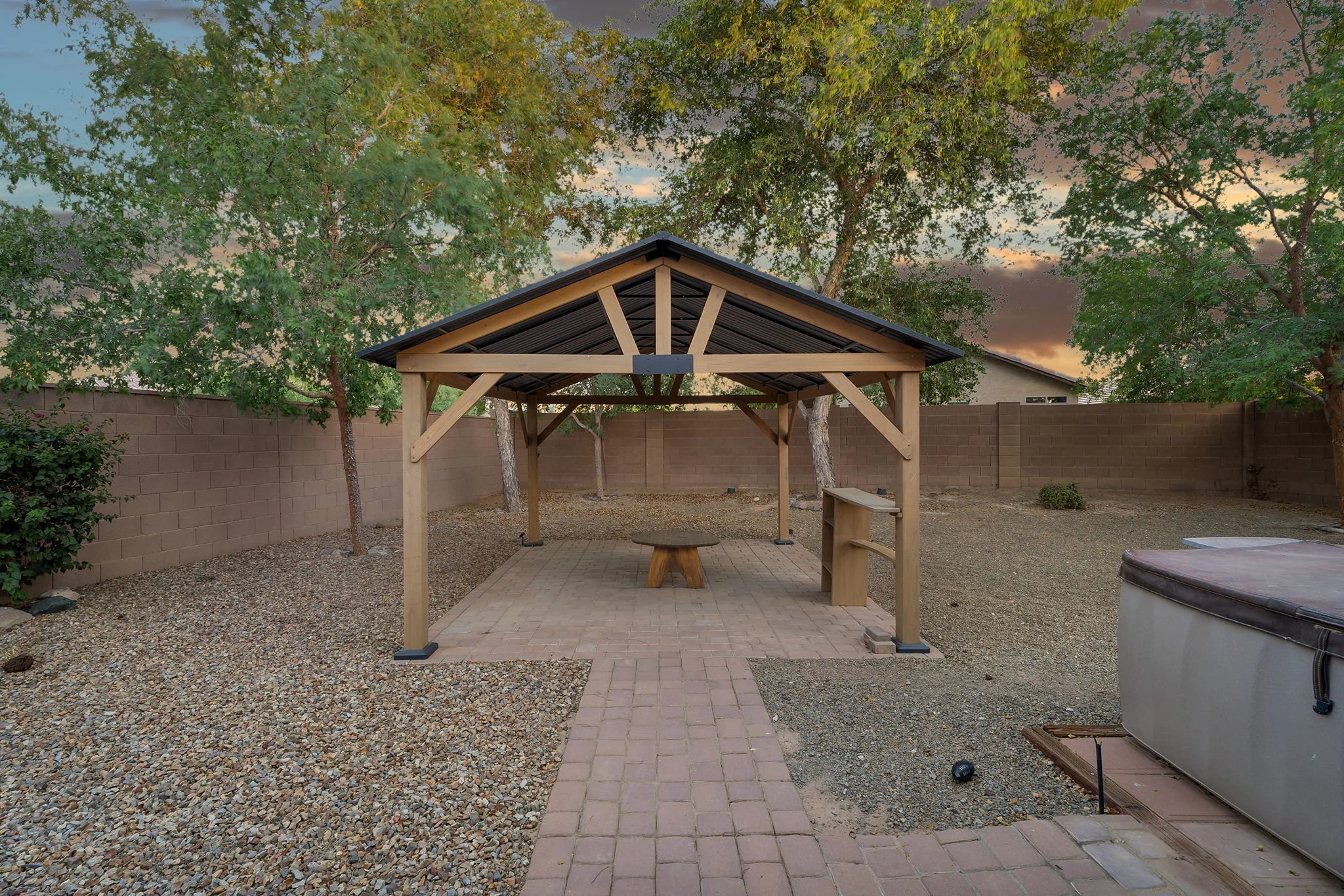 A backyard with a wood gazebo, brick pathway, gravel ground, trees, a brick wall, and some outdoor furniture at sunset.