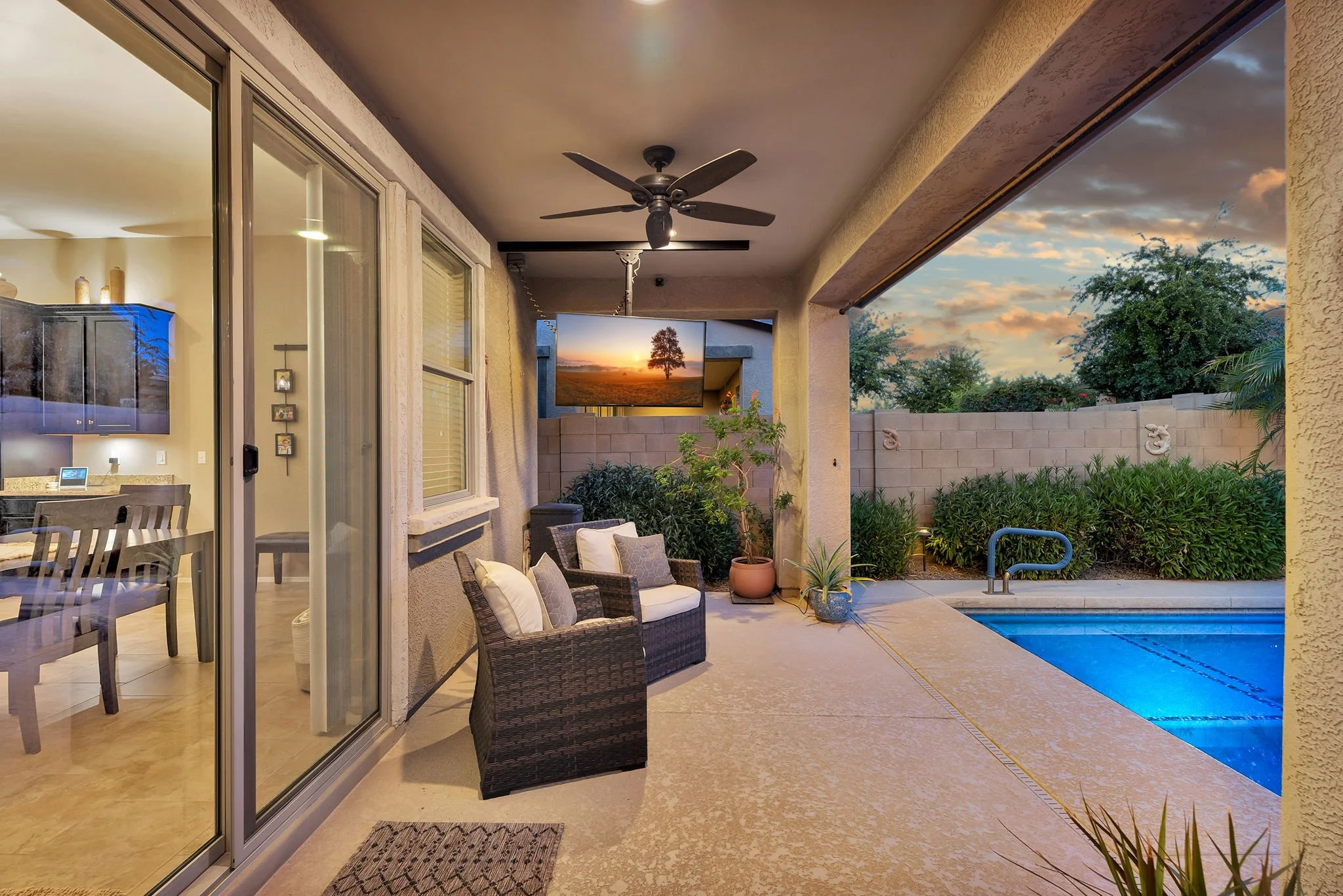 Covered patio area with outdoor seating, potted plants, and a view of the backyard with a swimming pool and trees during sunset.