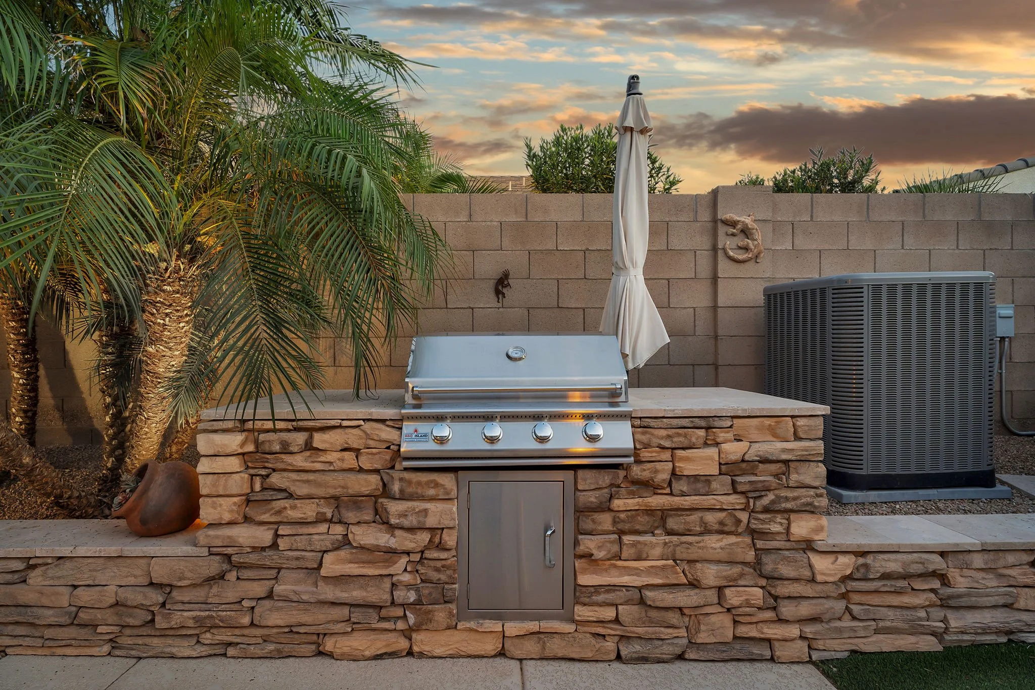 Outdoor backyard with a built-in grill, stone wall, and outdoor decor including a lizard sculpture, palm trees, a closed patio umbrella, and an air conditioning unit against a sunset sky with clouds.