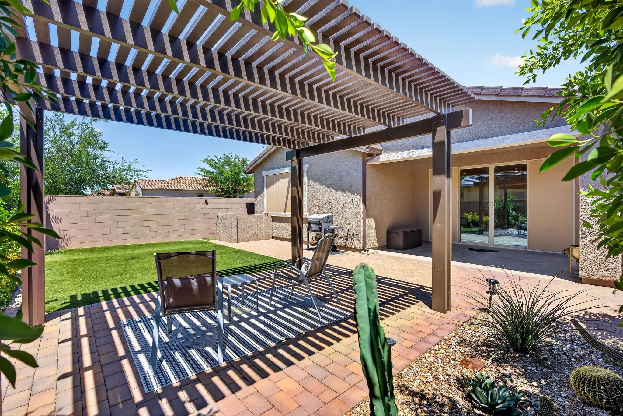 Backyard patio with a pergola providing shade, surrounded by green plants and a brick wall, with outdoor chairs and a grill near the house.