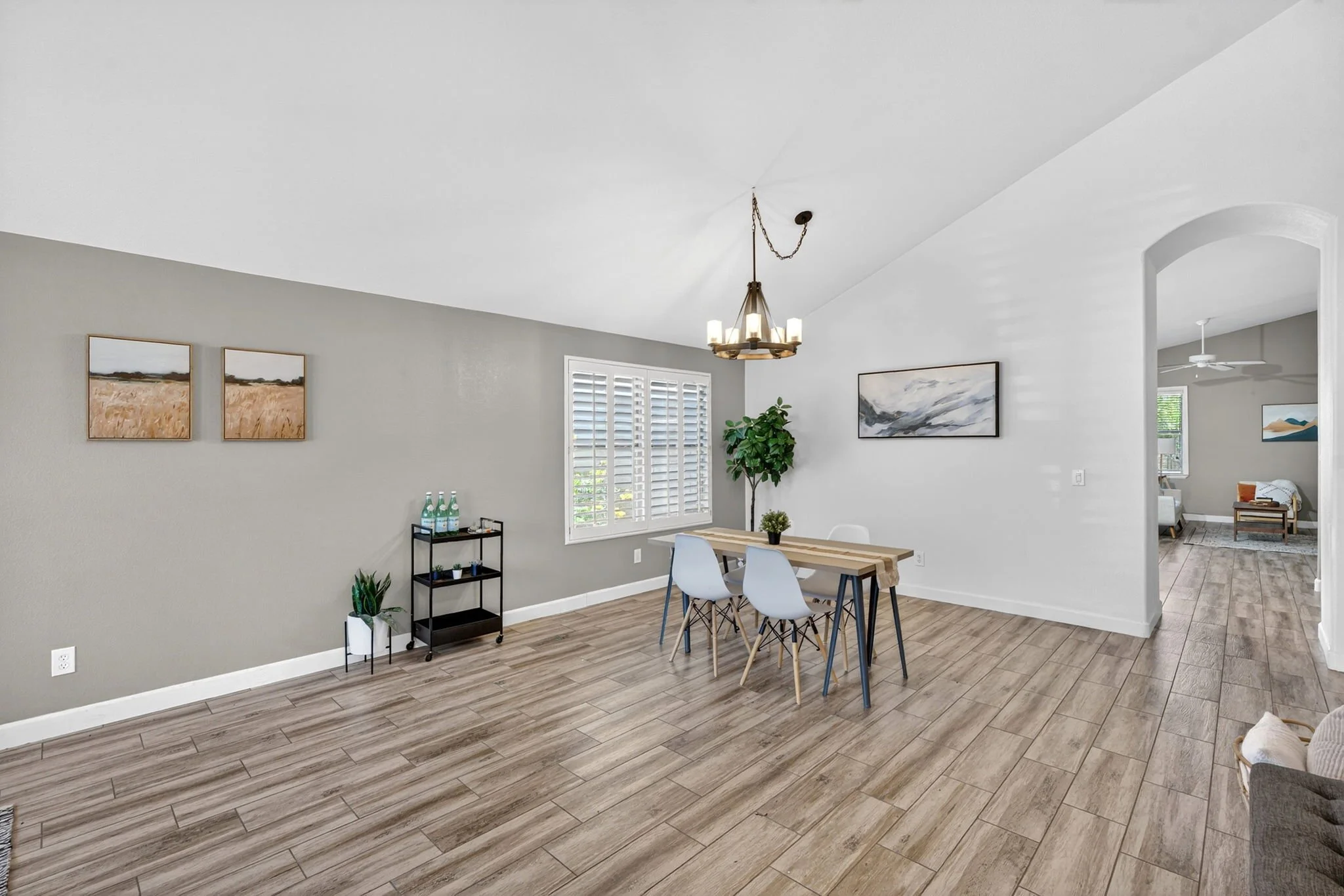 Interior of a modern living room with wooden floor, light-colored walls, a dining table with white chairs, wall art, a chandelier, and a large window with shutters.