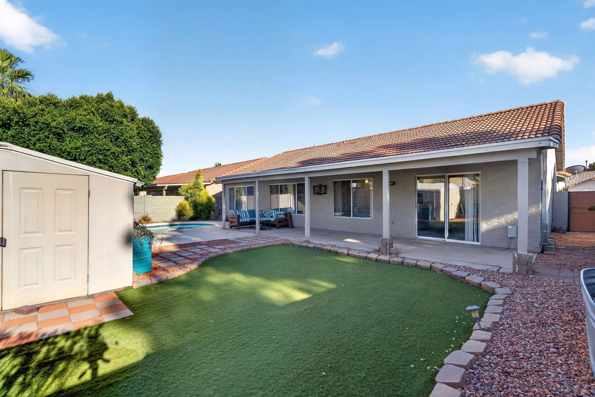 Backyard with grass lawn, patio area with outdoor furniture, house with beige walls and a tiled roof, shed, trees, and a clear blue sky.