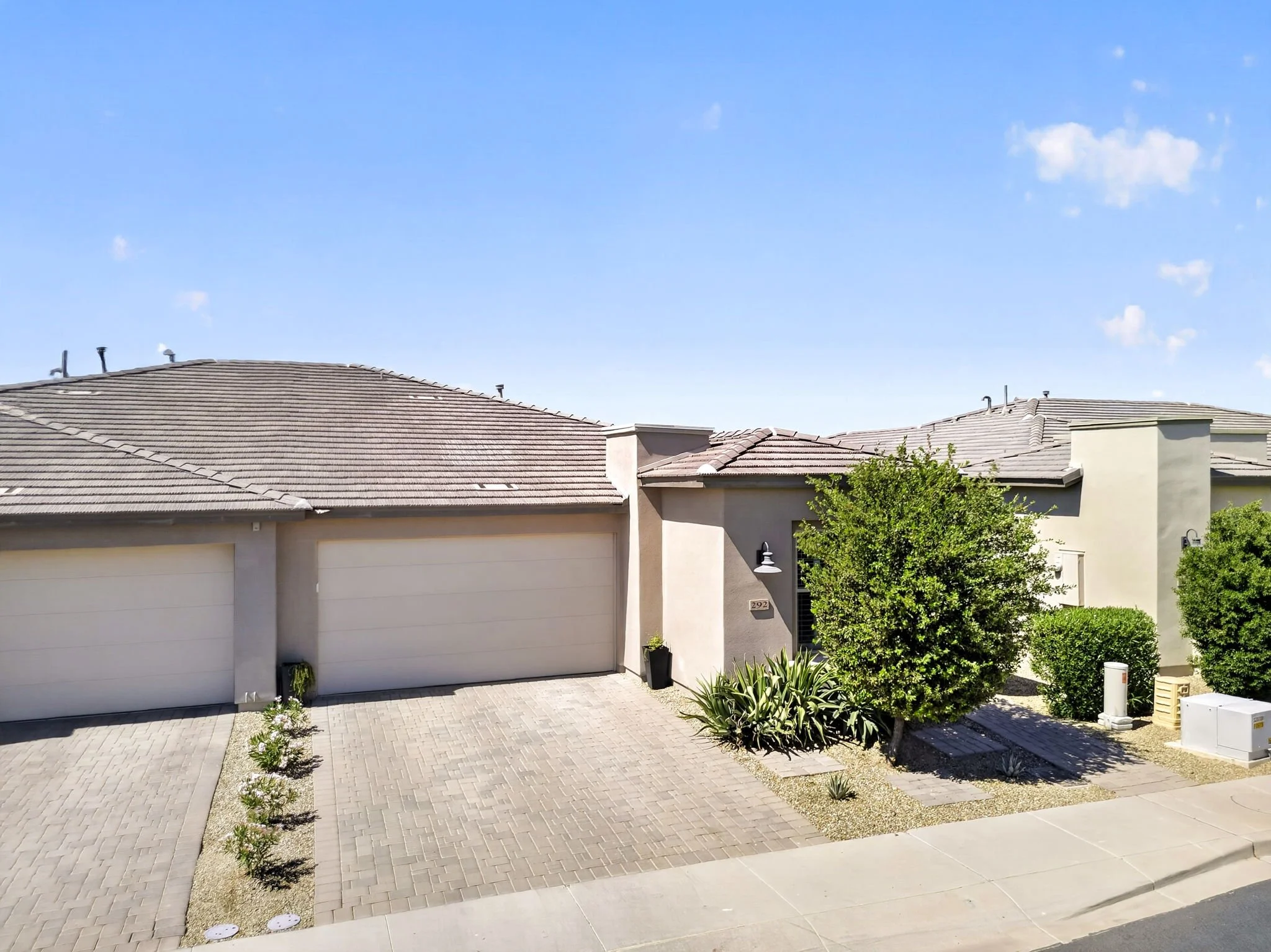 A residential house with a beige exterior and a tiled roof, two garage doors, a driveway paved with bricks, and a small garden with bushes and plants in front.
