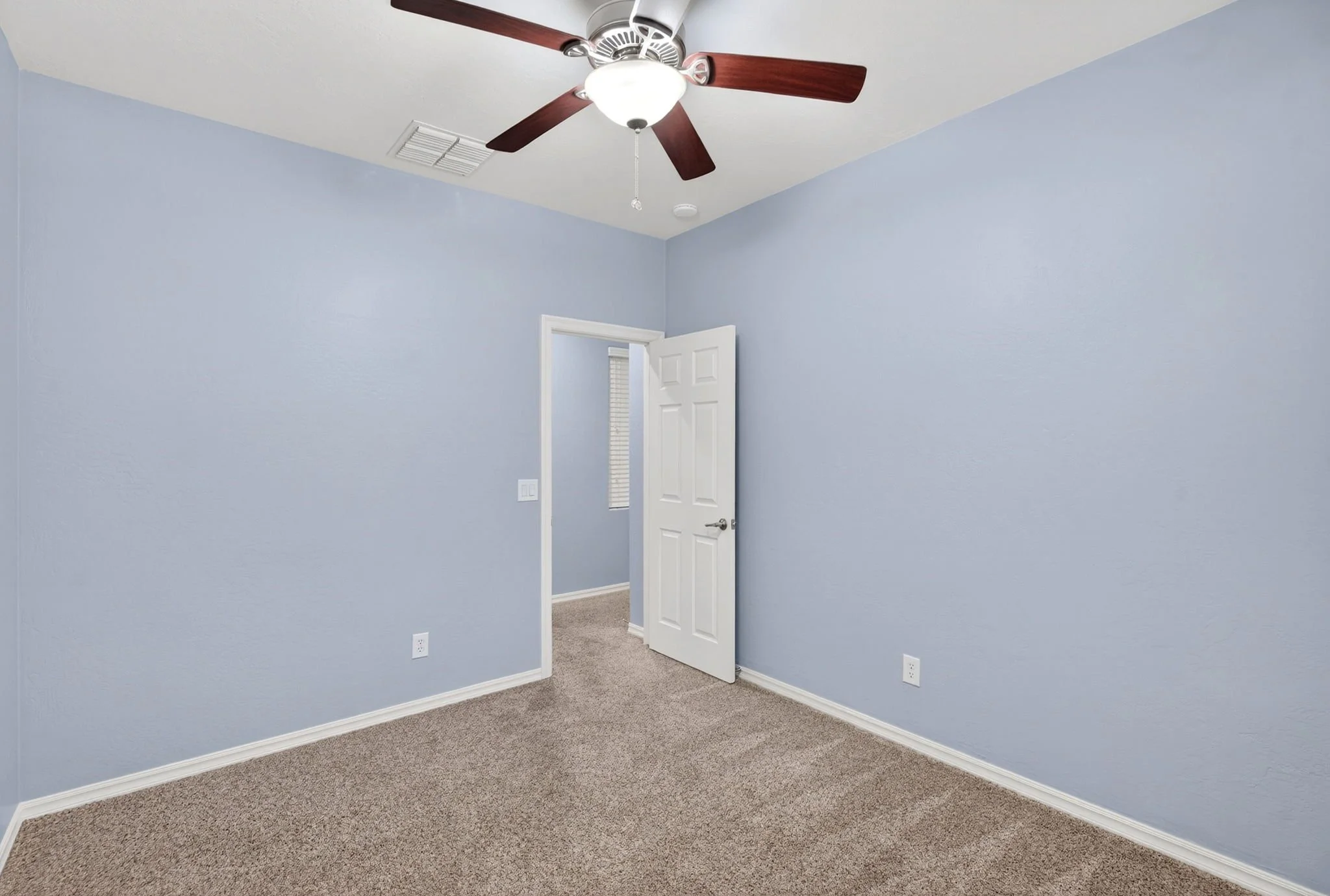An empty room with light blue walls, beige carpet, a white door slightly open, and a ceiling fan with wooden blades and a light fixture.
