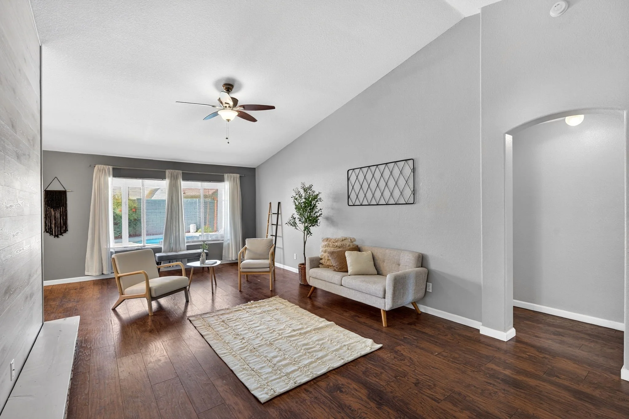 Living room with a ceiling fan, large window with white curtains, beige sofa, armchairs, a ladder, plants, and a beige rug on wooden floor.