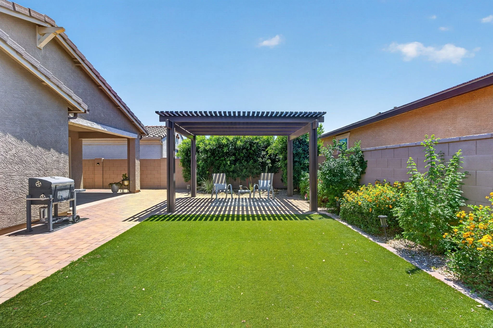 A backyard with a lawn, paver patio, and a wooden pergola. There are surrounding houses with beige and gray exterior walls, a grill on the patio, and flowering bushes along the fence. Two chairs are placed under the pergola, and the sky is clear with