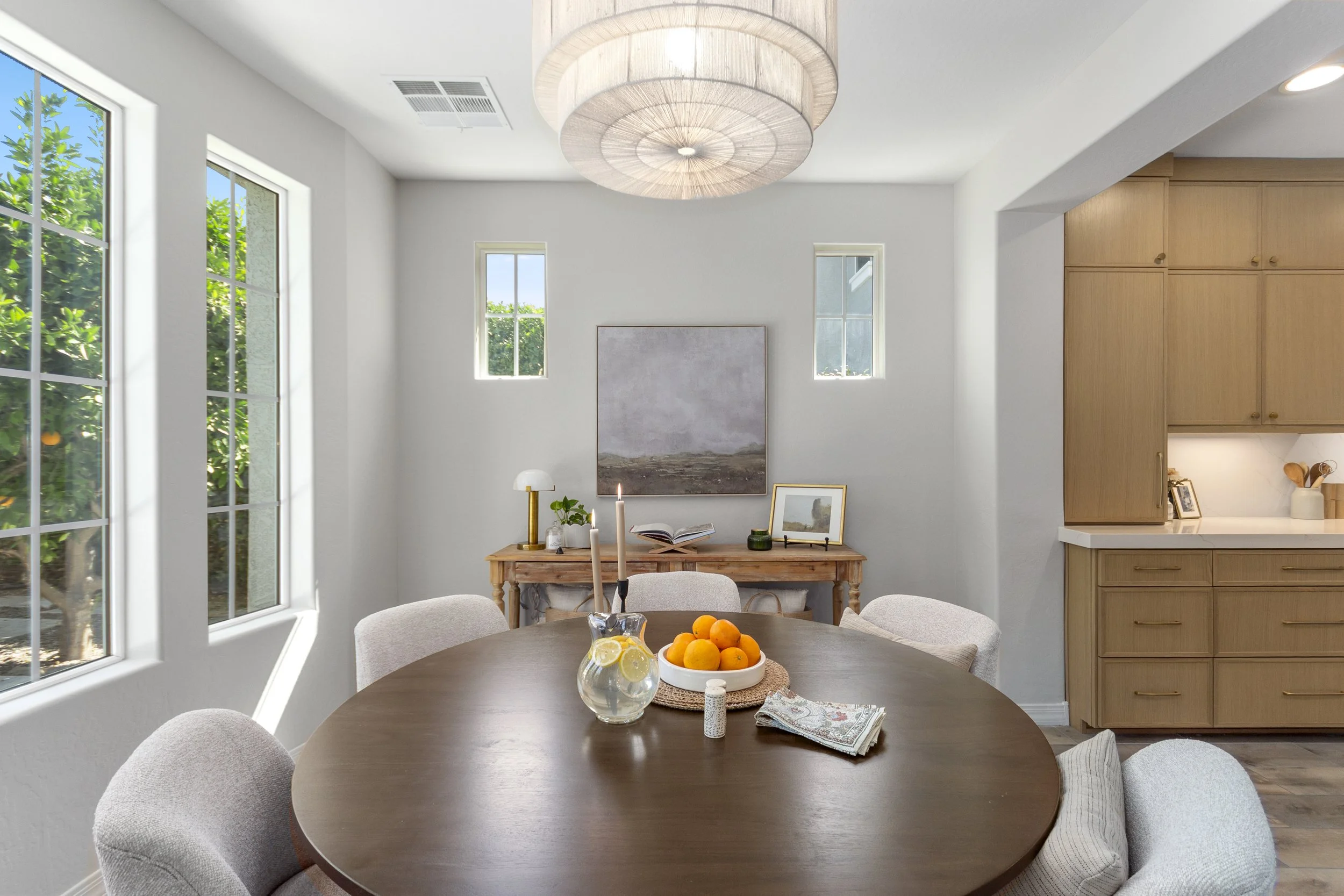 A dining room with a round wooden table surrounded by beige upholstered chairs. On the table is a glass pitcher of lemonade with lemon slices, a bowl of oranges, and a decorative cloth. The room has large windows with greenery outside, white walls, a