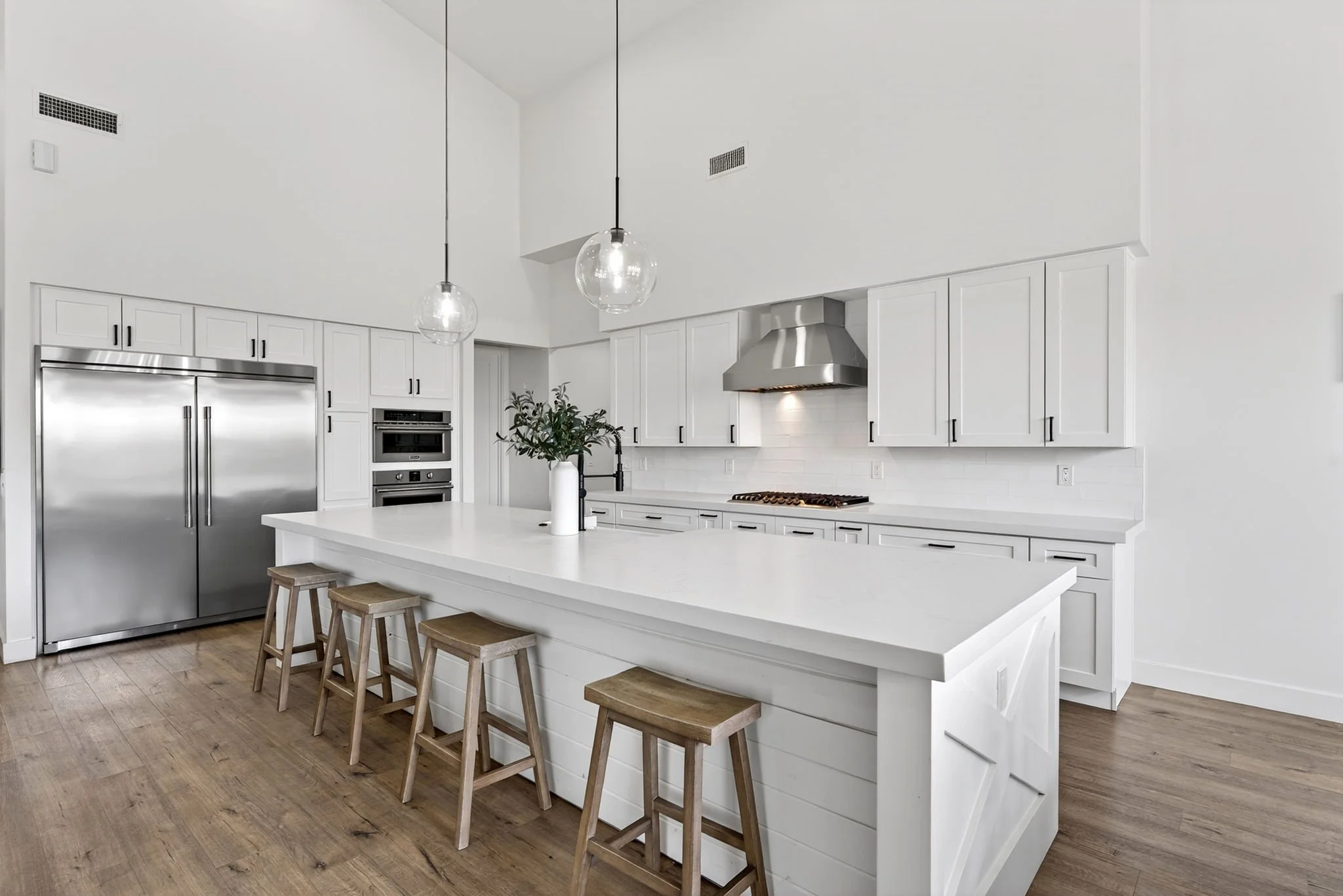 A modern, white kitchen with a large island, stainless steel refrigerator, built-in oven, and a range hood. There are three wooden stools at the island, a small plant in a white vase, and pendant lights hanging from the ceiling.