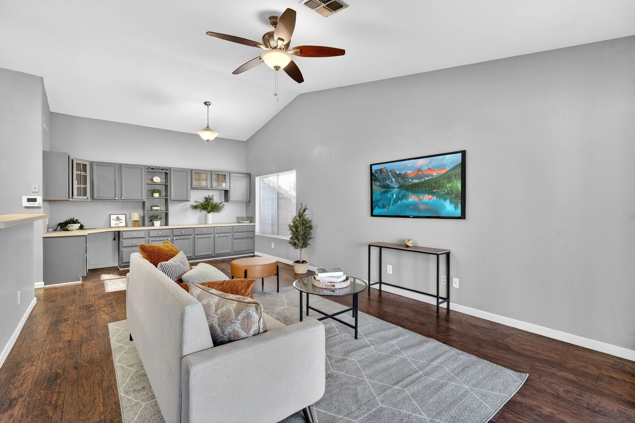 Living room with gray walls, ceiling fan with wooden blades, mounted flat-screen TV displaying a mountain and lake scene, gray cabinetry, with a couch, coffee table, and decorative indoor plants.