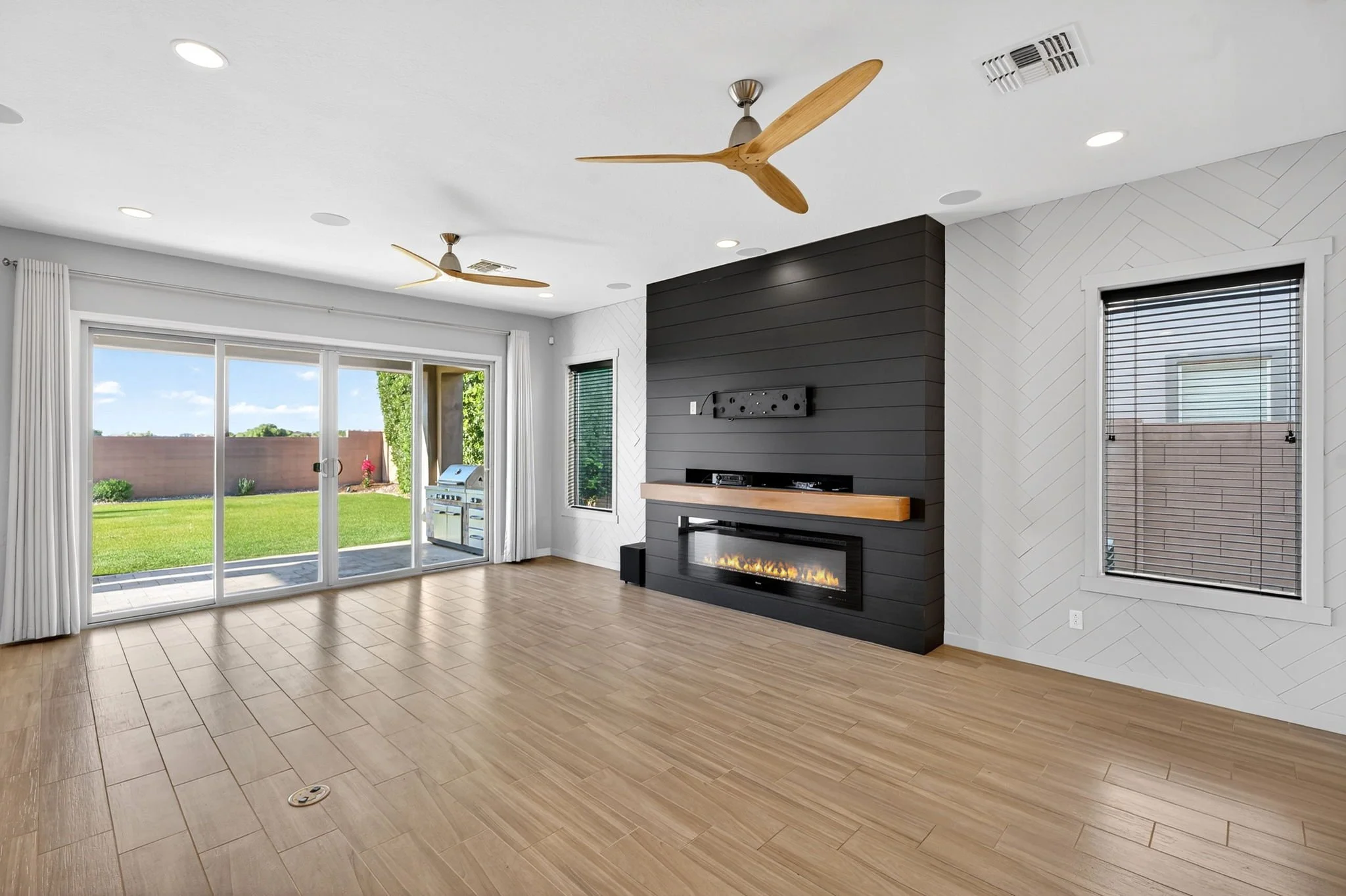 A modern living room with a large glass sliding door opening to a backyard with green grass and a fence. The room features wood-look tile flooring, a black accent wall with a built-in fireplace, a wall-mounted TV, and two ceiling fans with wooden bla