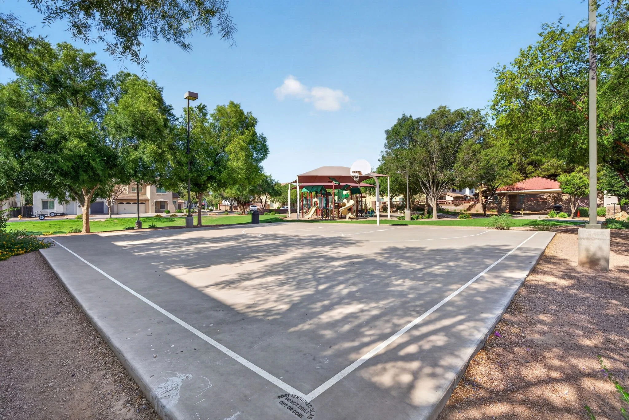 An empty outdoor basketball court with a playground in the background, surrounded by trees and residential houses under a clear blue sky.