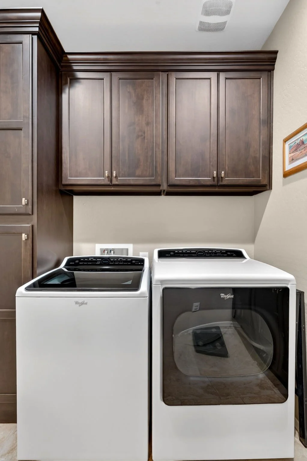 A laundry room with a top-loading washer on the left and a front-loading dryer on the right, both white, with dark control panels at the back. Overhead, there are wooden cabinets with simple panel doors and small knobs.