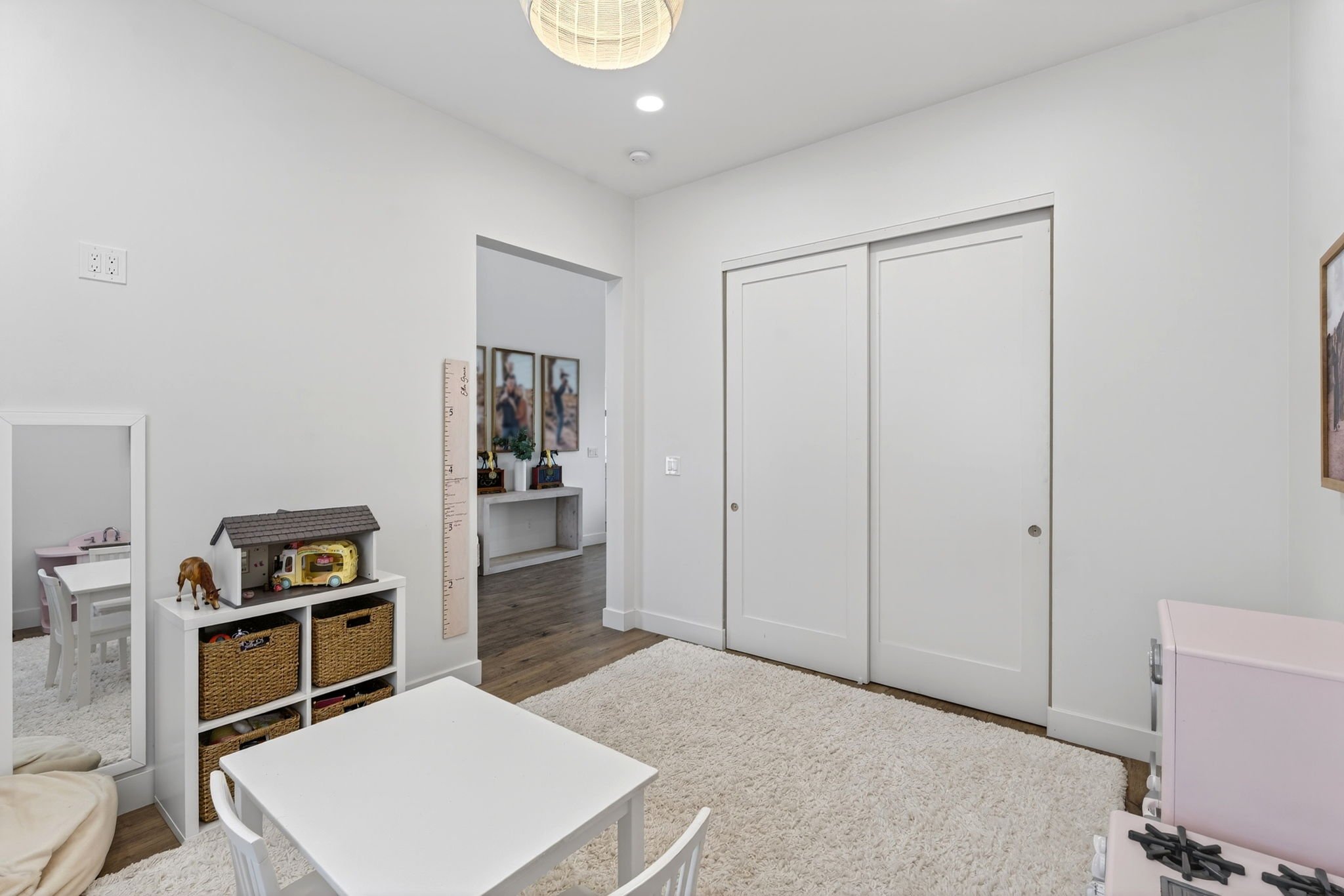 A child's play area with white furniture, wicker baskets, and toys, next to a closet door and wall decor, with beige carpet and hardwood flooring.
