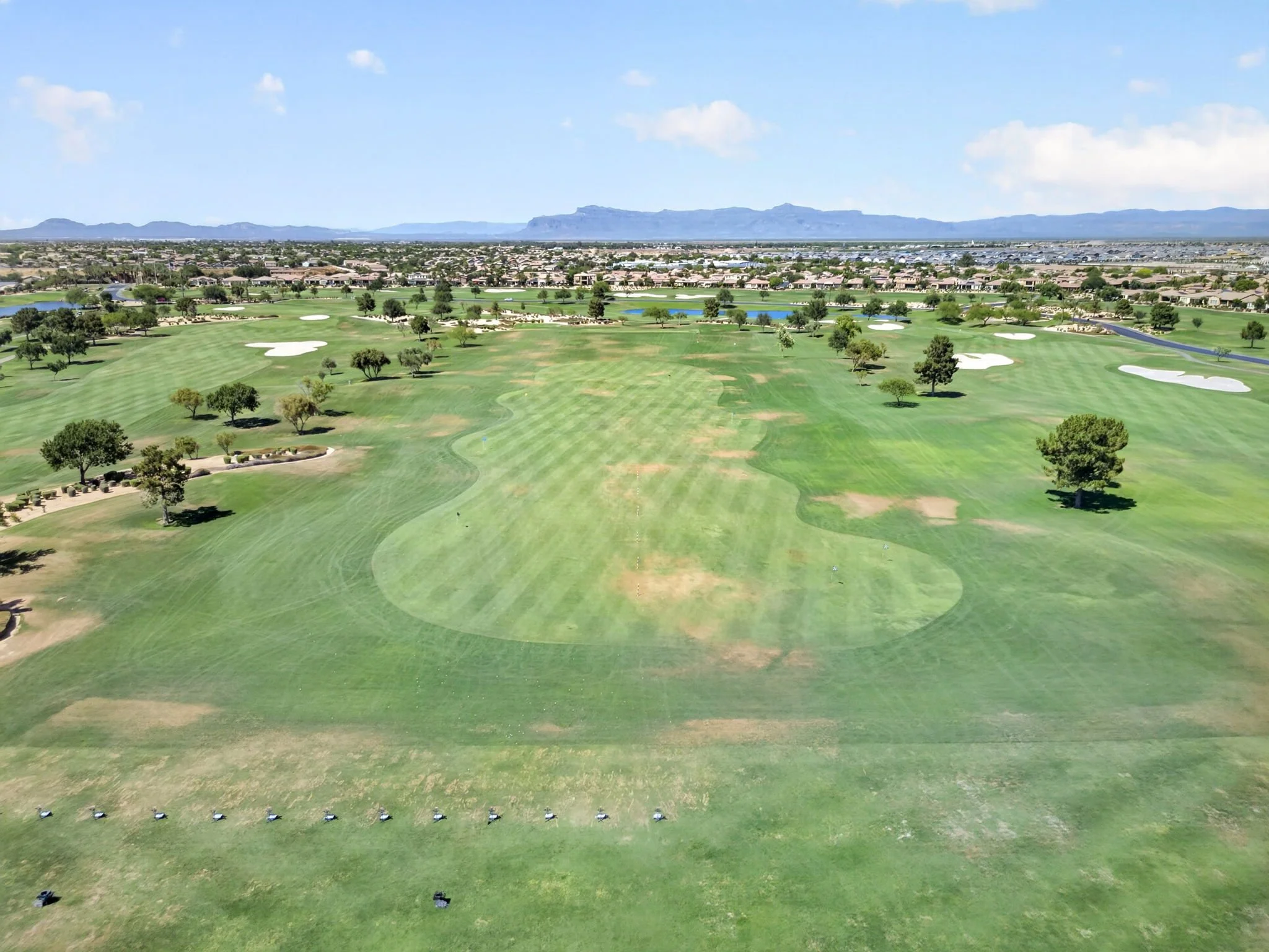 A view of a golf course with green fairways, sand traps, and water hazards, surrounded by trees and residential houses, with mountains in the distance under a blue sky with a few clouds.