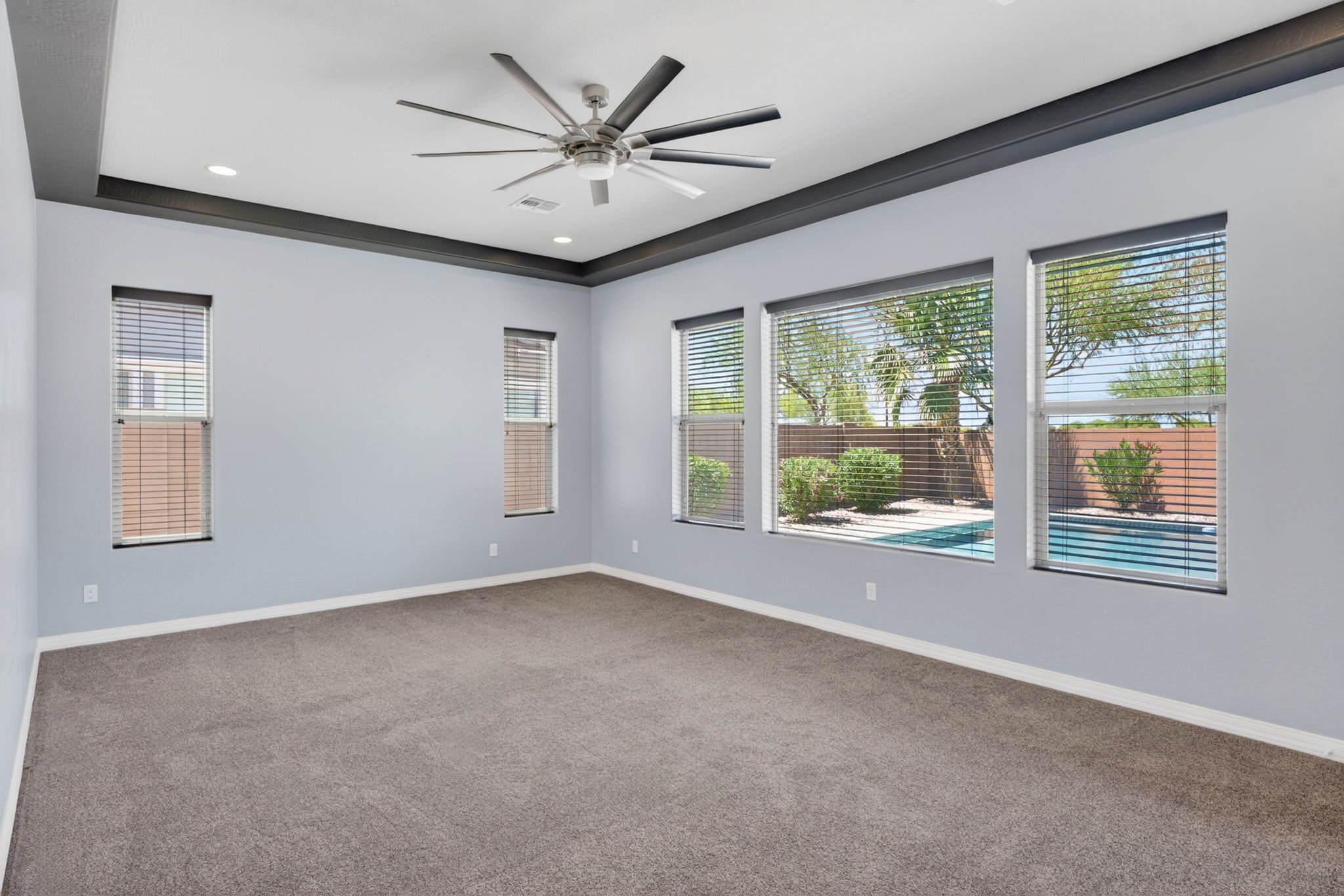 Empty living room with large windows, ceiling fan, gray walls, beige carpet, and outdoor view of trees and a pool.
