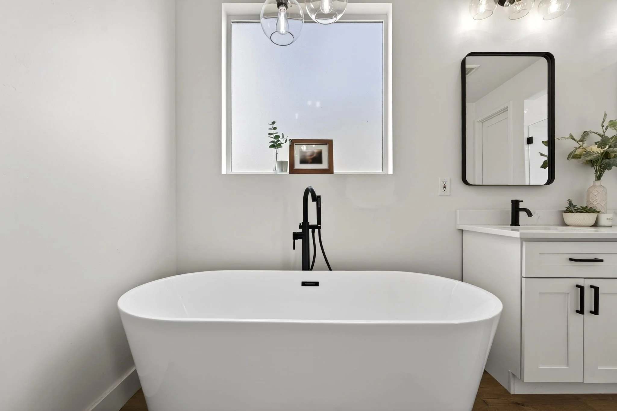 Modern bathroom featuring a white freestanding bathtub, a black floor-mounted faucet, decor with potted plants, a framed photograph, a mirror with a black frame, and a large frosted window with hanging glass light fixtures.