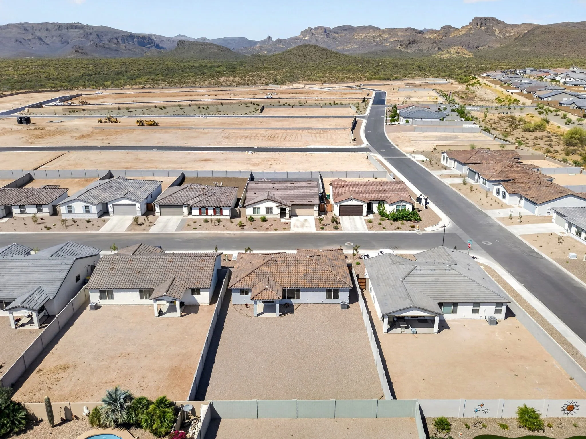 A row of new houses under construction in a suburban neighborhood, with dirt lots and construction machinery, set against arid mountainous landscape in the background.